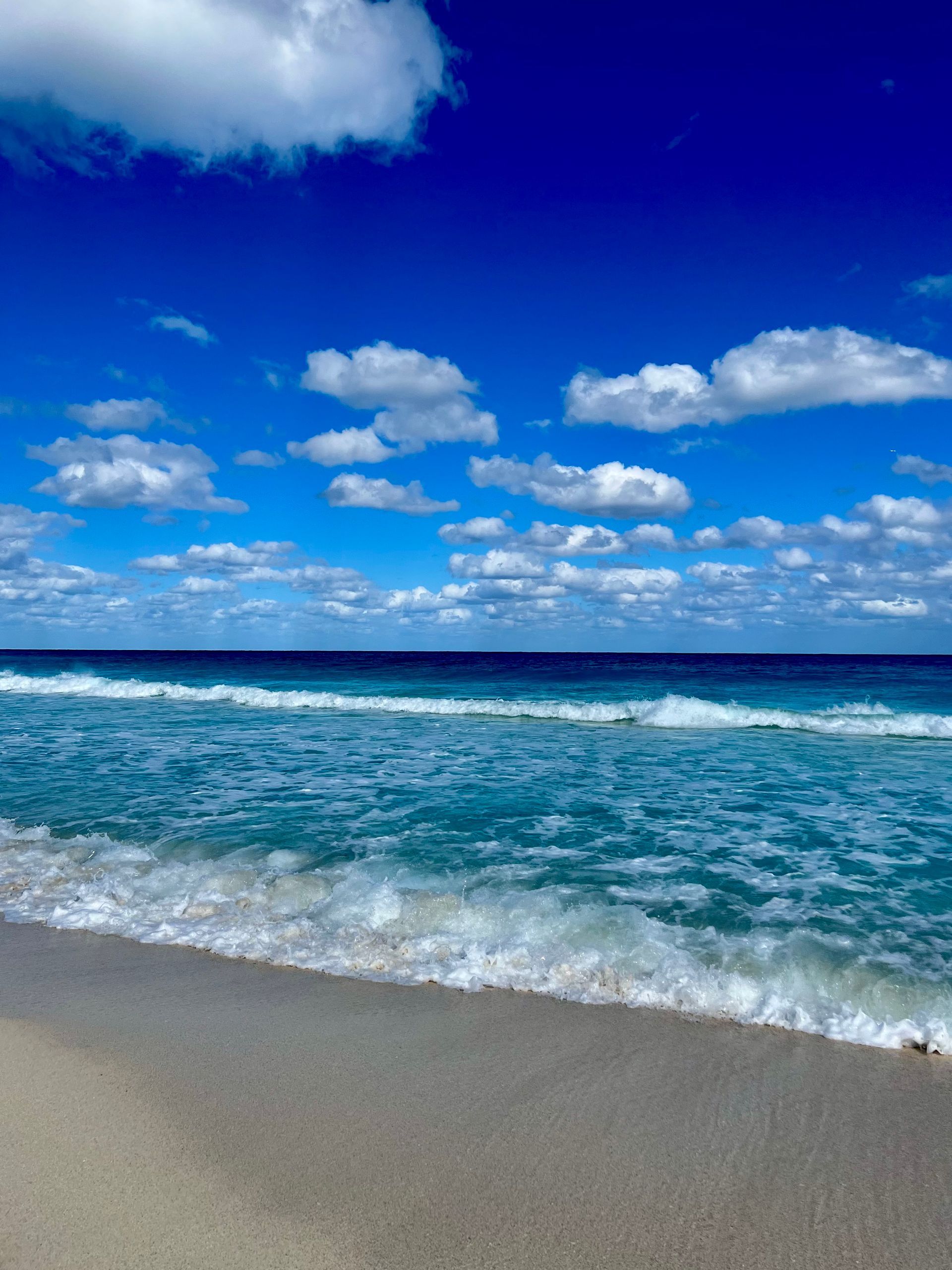 Las olas del océano bañan suavemente una playa de arena bajo un cielo azul brillante con nubes blancas dispersas.