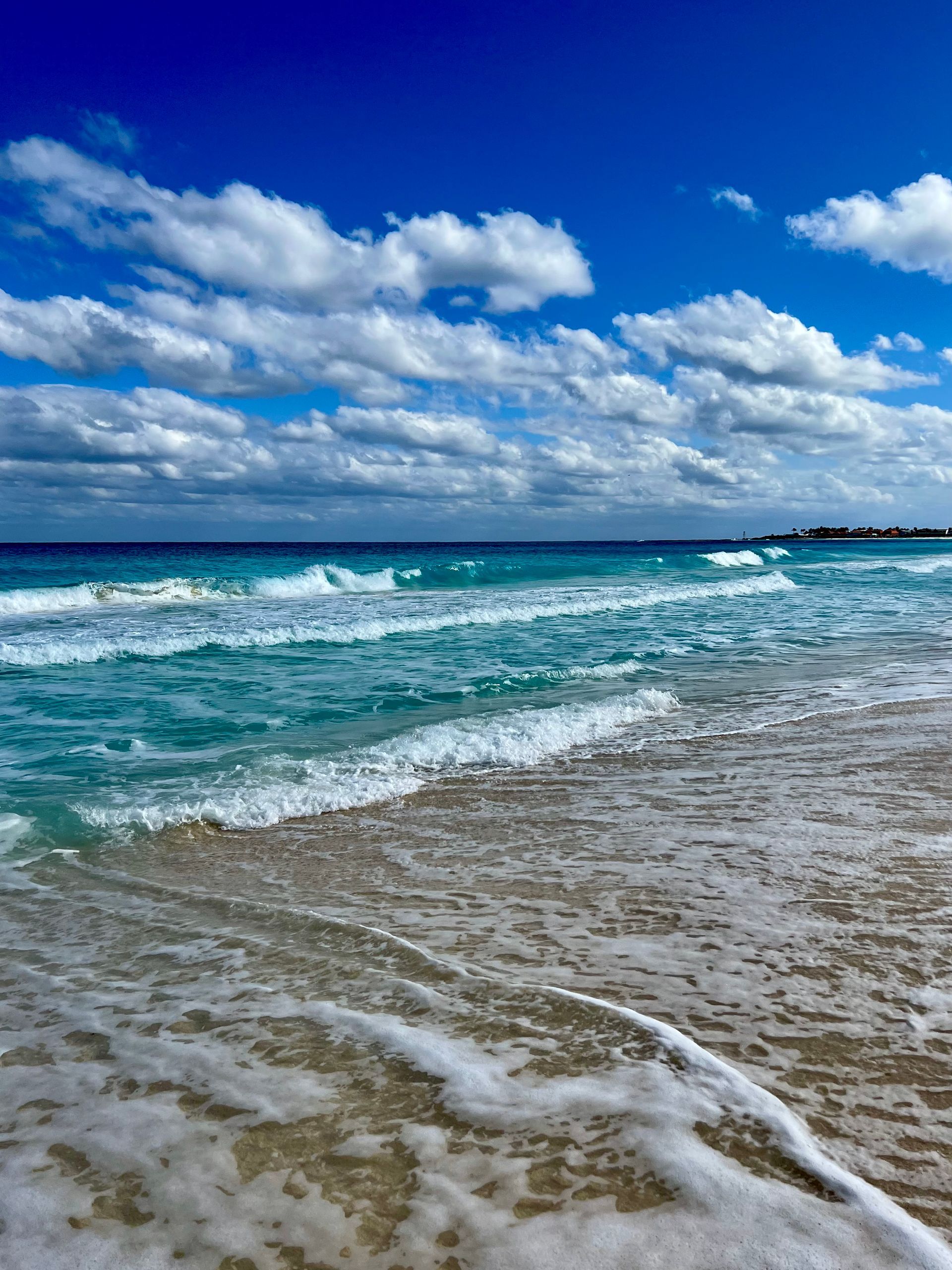 Olas del océano rompiendo en una playa de arena bajo un cielo azul brillante con nubes blancas.