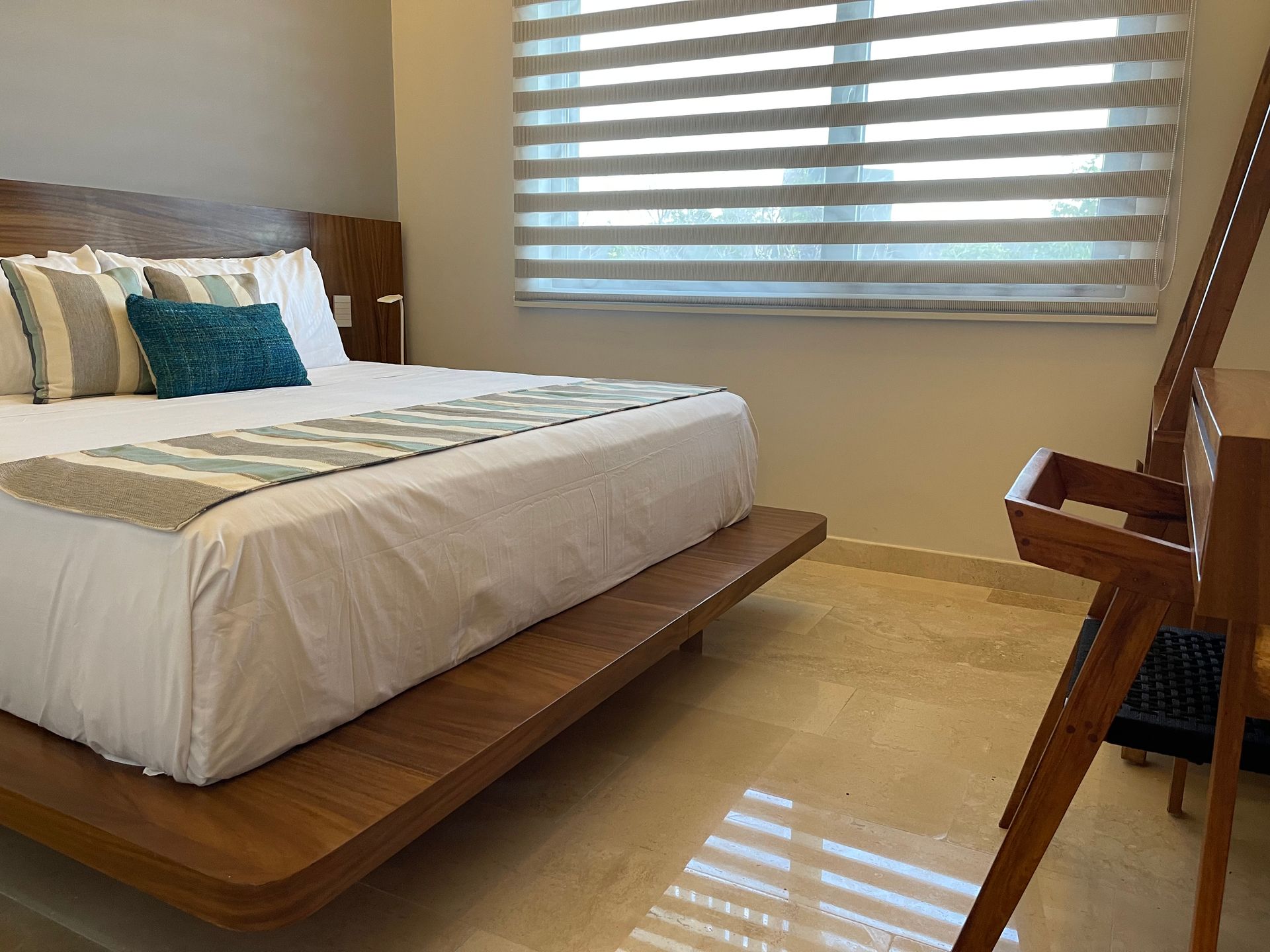 Dormitorio con cama de madera, ropa de cama de color claro, almohadas y ventana con persianas.