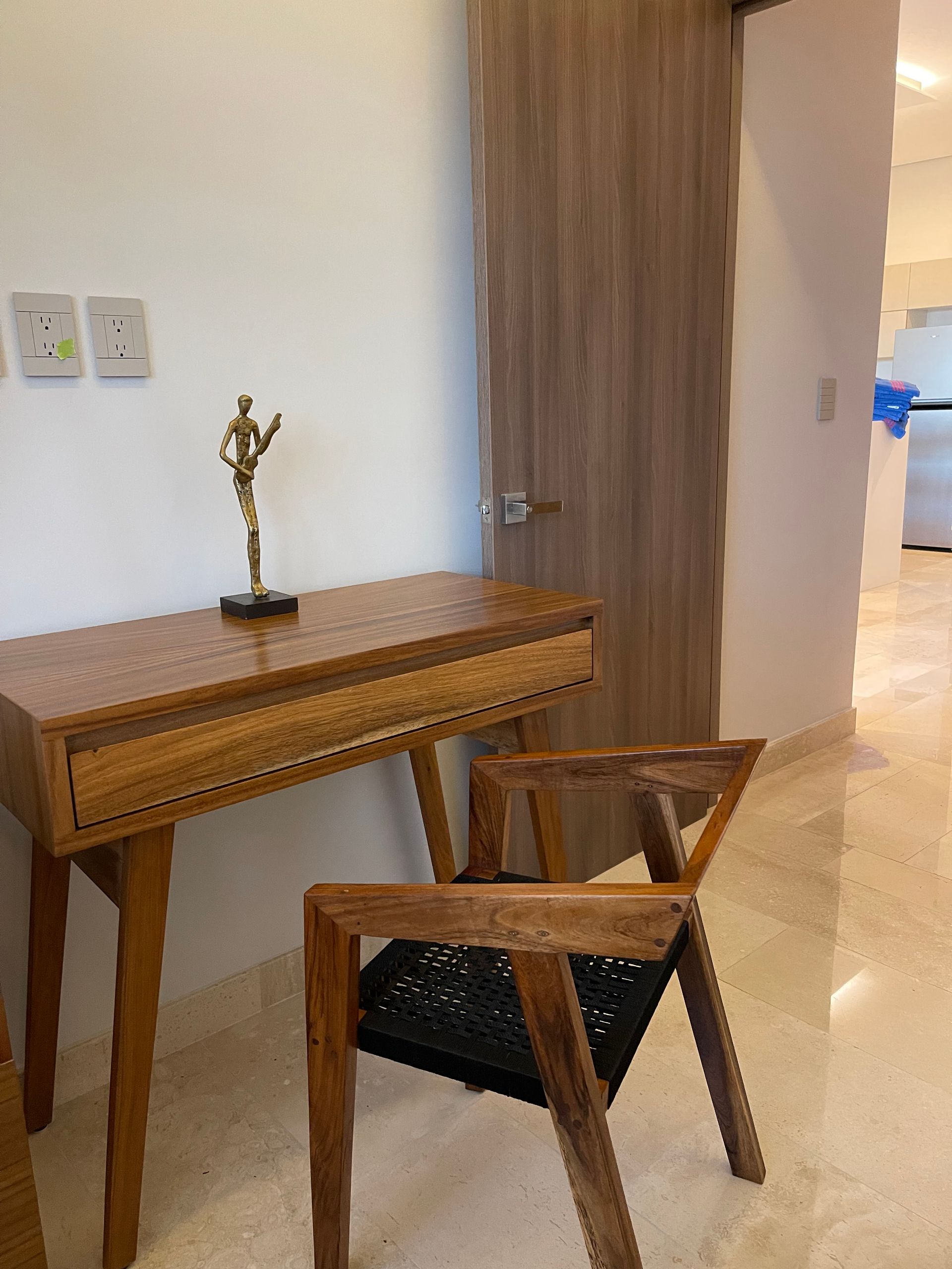 Escritorio de madera con estatua, silla  junto a puerta cerrada en una habitación con paredes y suelos de colores claros.