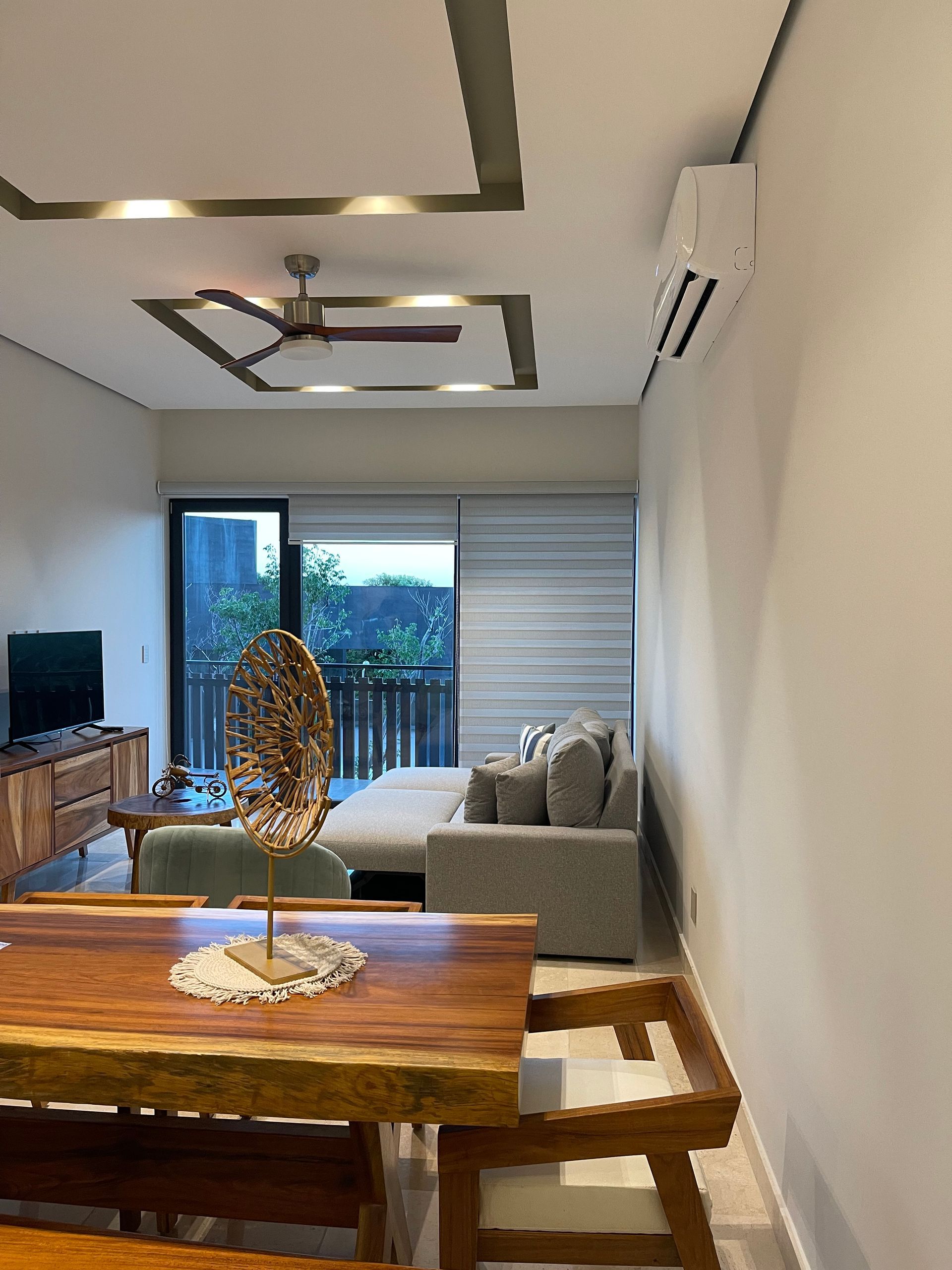 Sala de estar con muebles de madera, sofá, TV y acceso al balcón. Paredes y techo beige con iluminación empotrada.