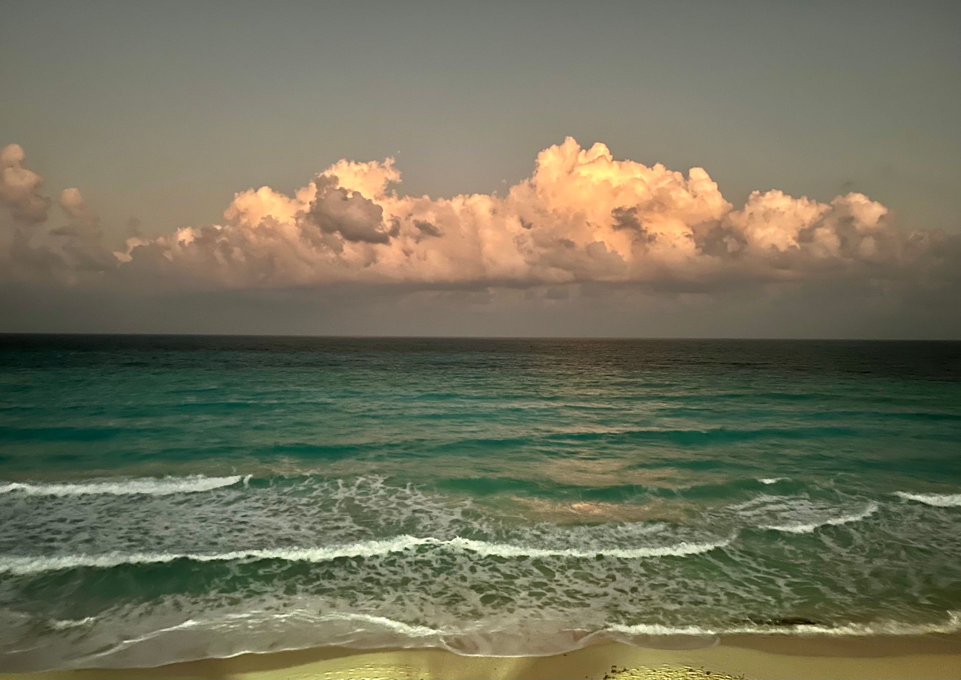 Ocean with turquoise water and waves meeting a sandy shore under a cloudy sky.