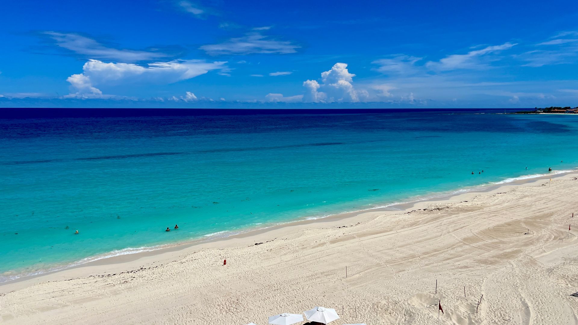 Playa de arena blanca, agua turquesa y cielo azul con nubes. Hay gente nadando.
