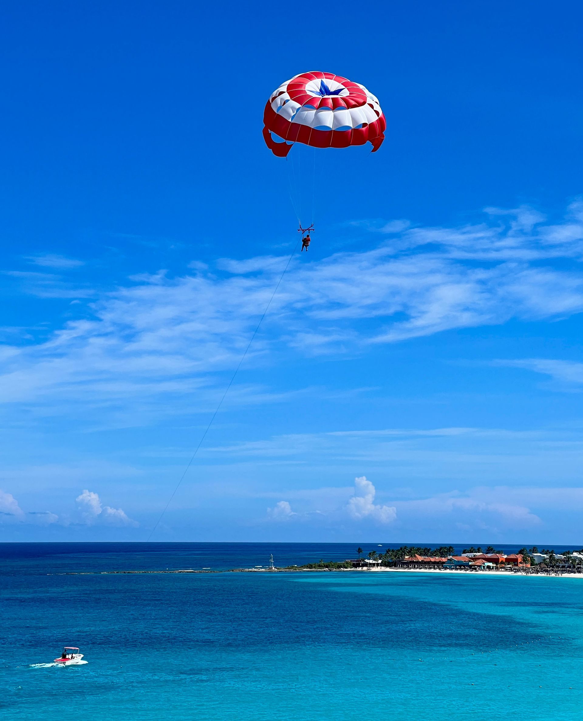 Parasailer in the air over turquoise ocean, blue sky. Small boat below.