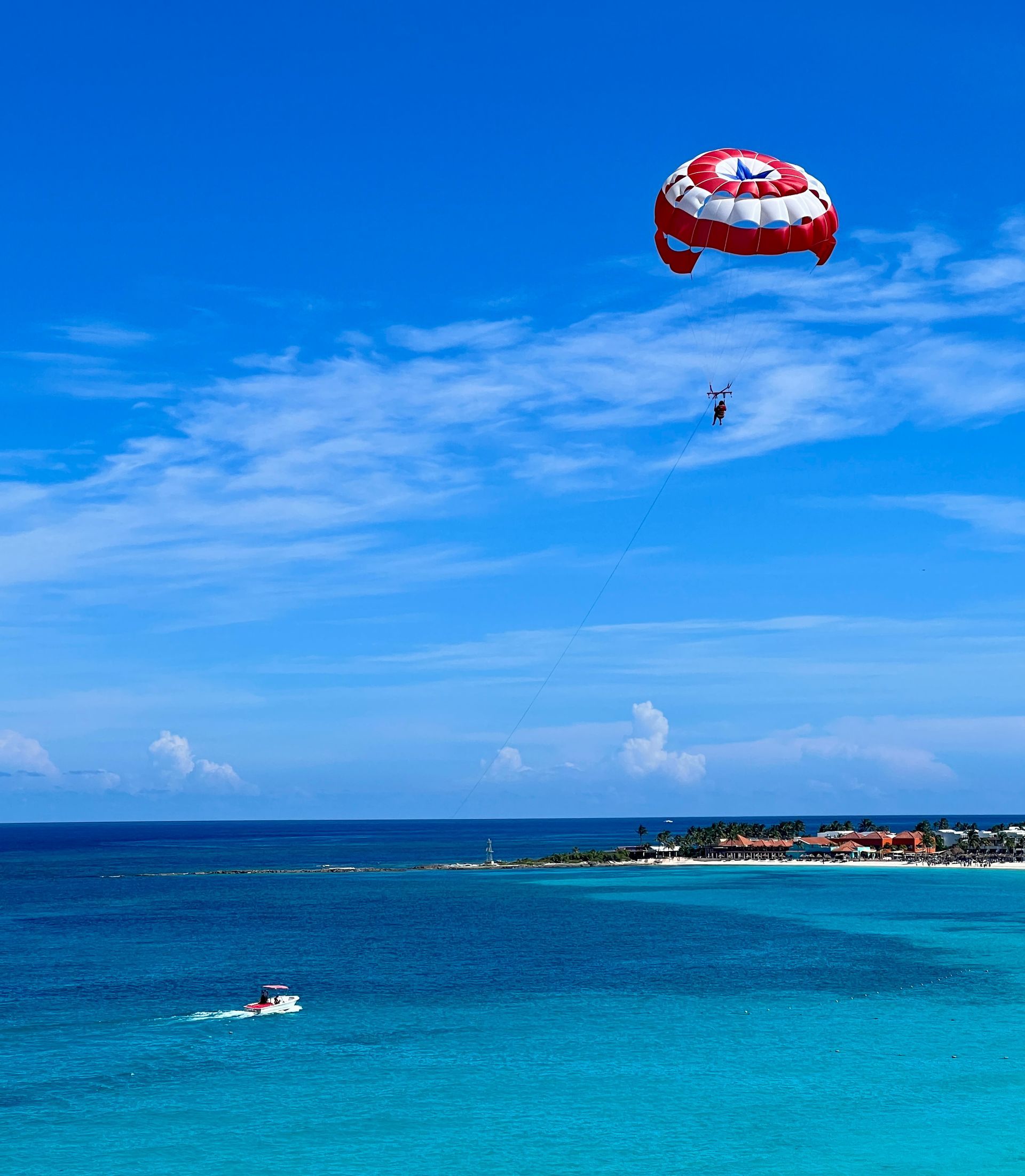 Parasailer high over turquoise ocean; white and red parachute, boat below.