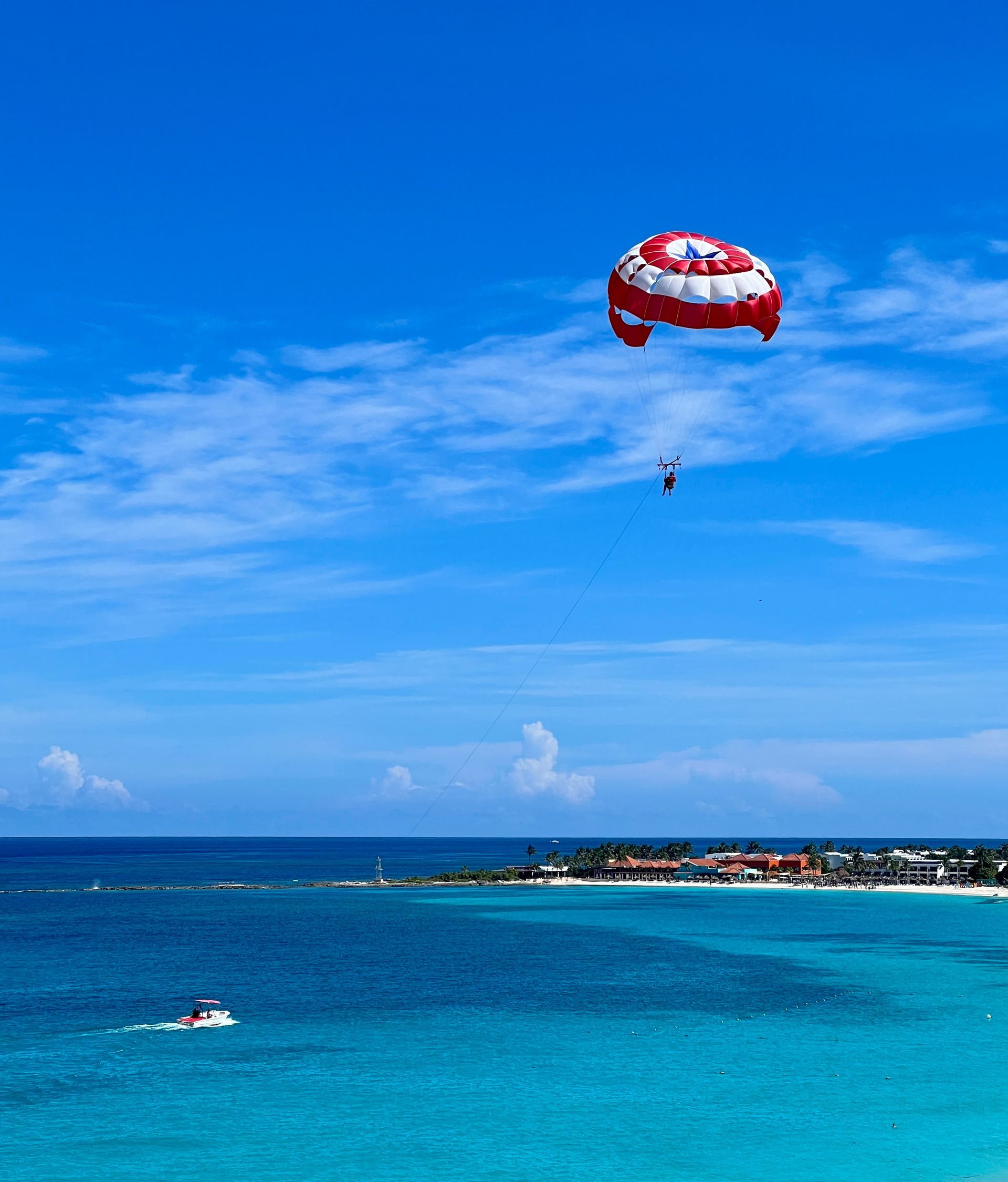 Parasailing over turquoise water; red and white parachute, sunny beach day.