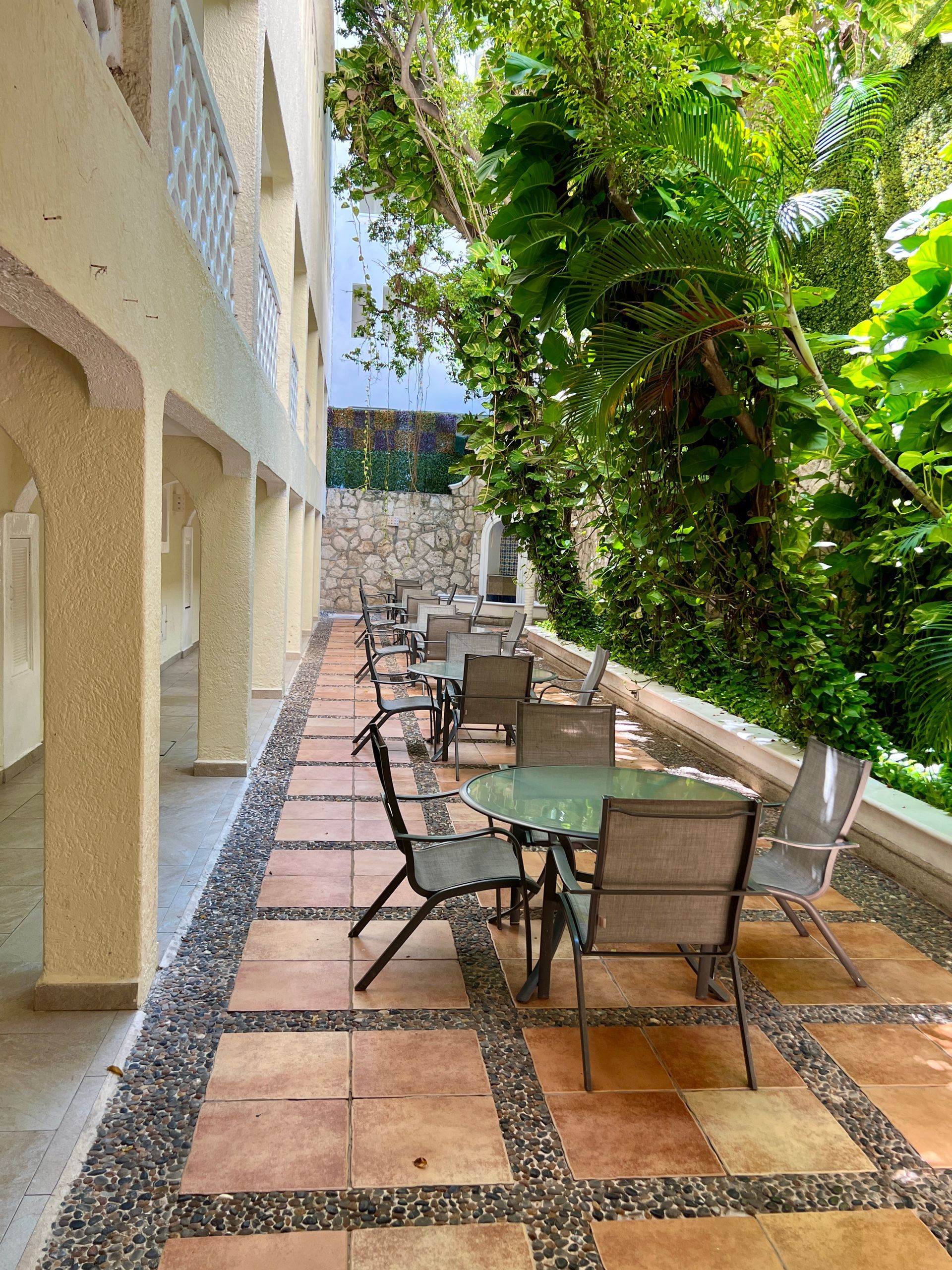 Outdoor dining area with tables and chairs under a building's covered walkway, next to lush greenery.