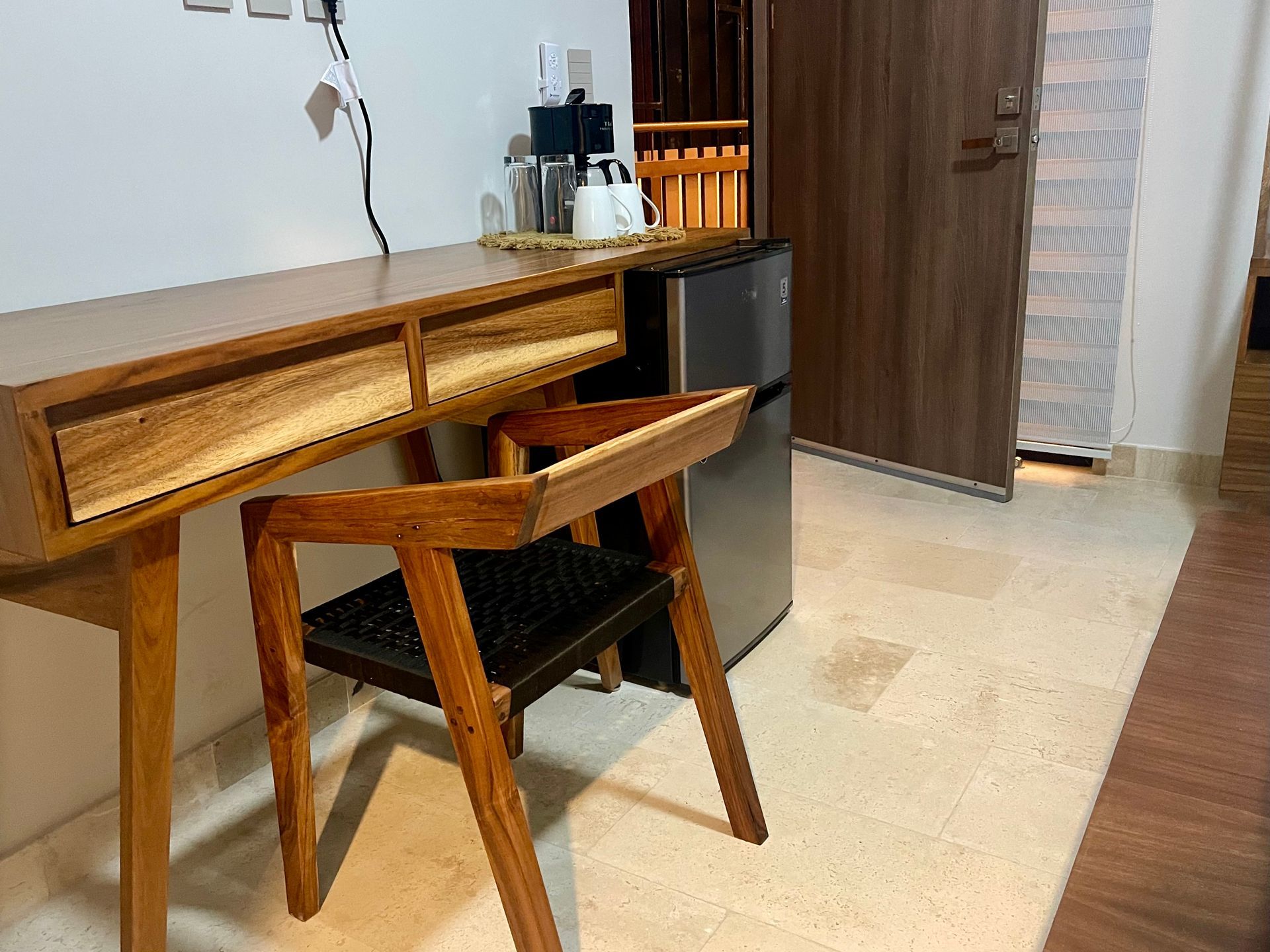 Escritorio de madera y silla junto a una mininevera y una puerta. Cafetera y tetera sobre el escritorio.
