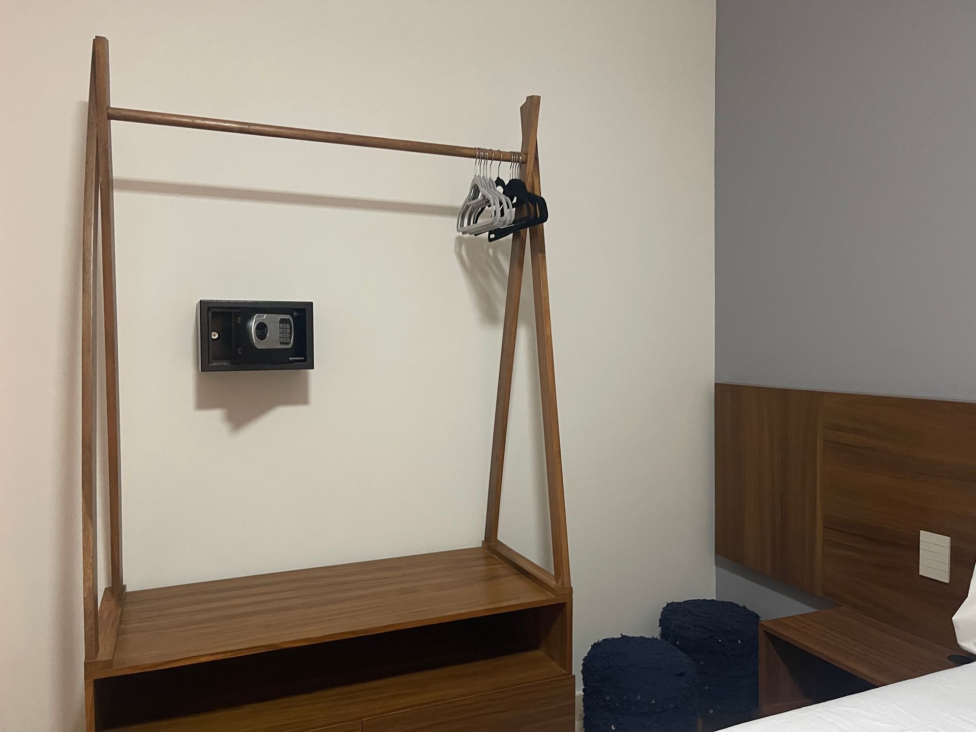 Perchero de madera con caja fuerte en la pared. Pequeños taburetes azules y una mesita de noche.