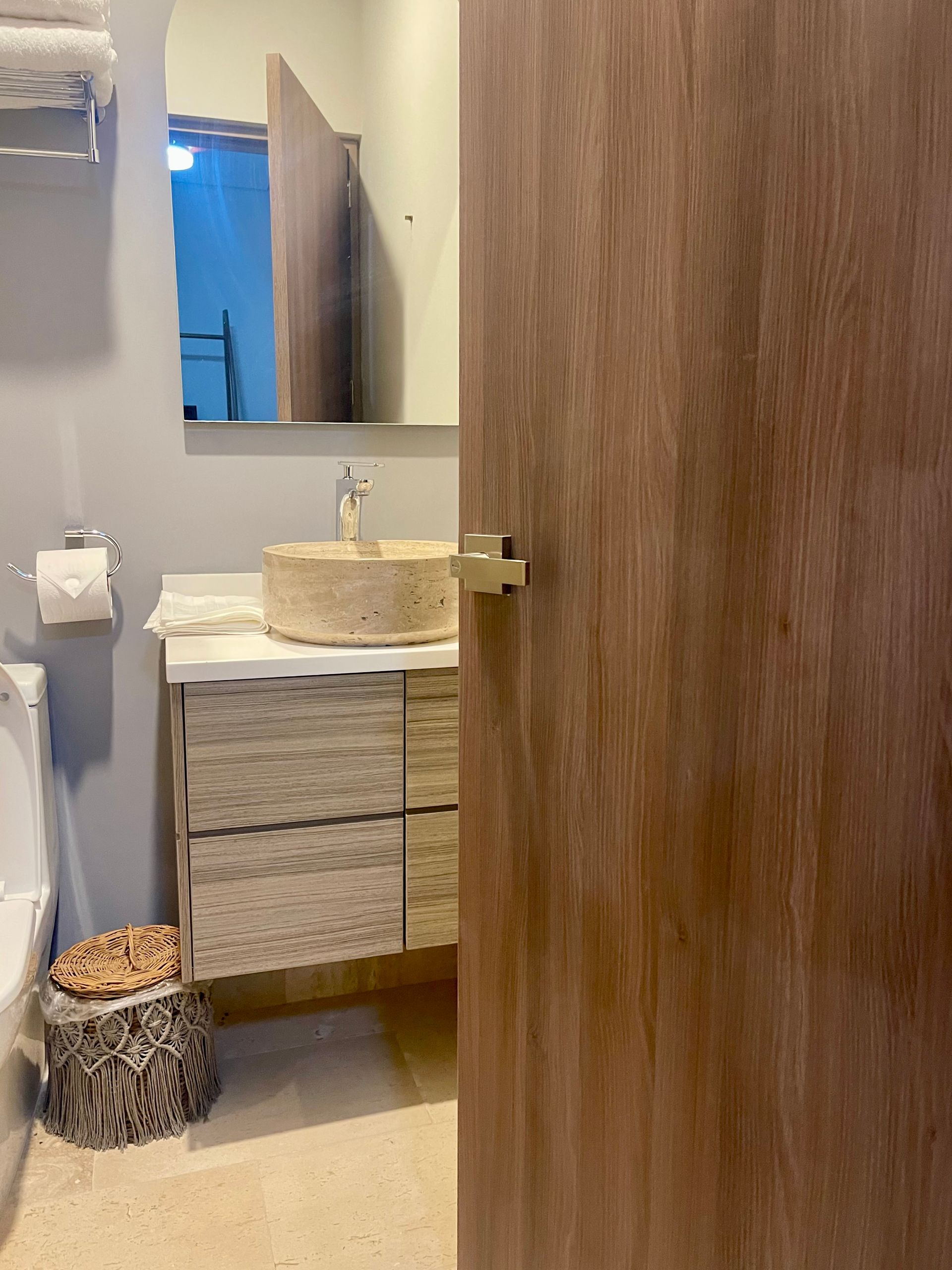 Baño con puerta de madera clara, tocador con lavabo de piedra, espejo e inodoro.