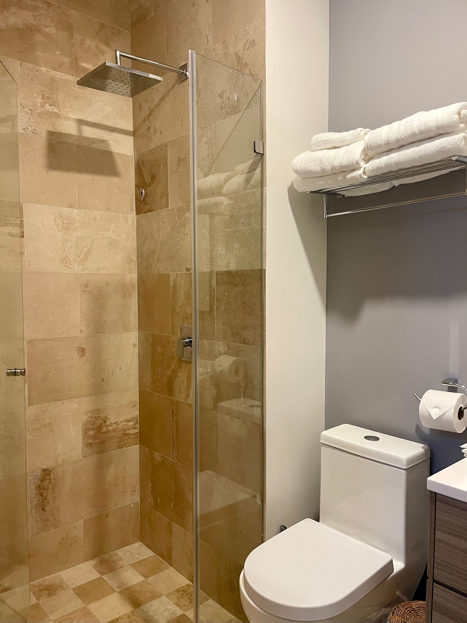Ducha con puerta de cristal, azulejos de color beige, inodoro y toallas en un estante en un baño con paredes grises.