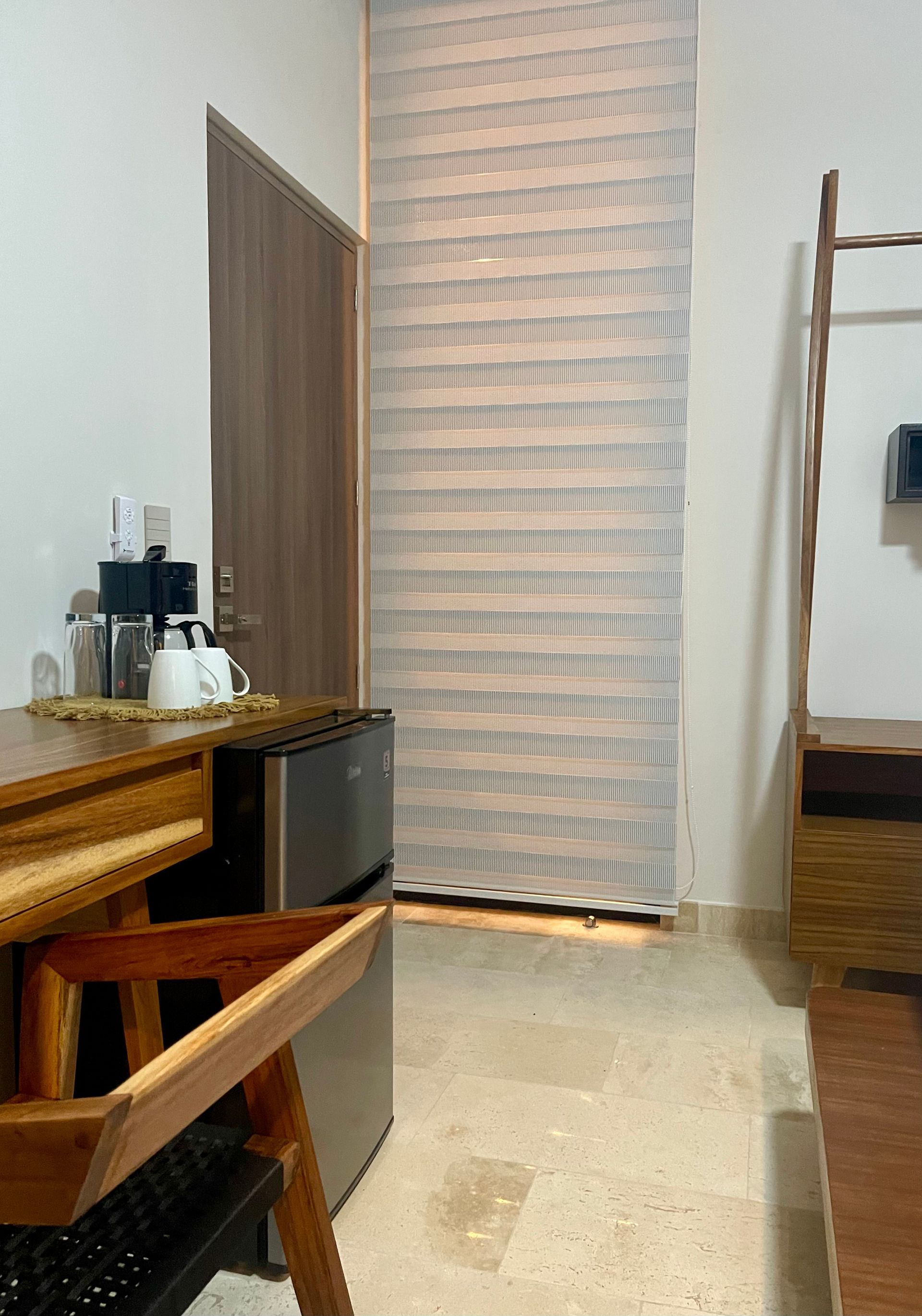 Una habitación pequeña con escritorio, refrigerador y una ventana con persianas.