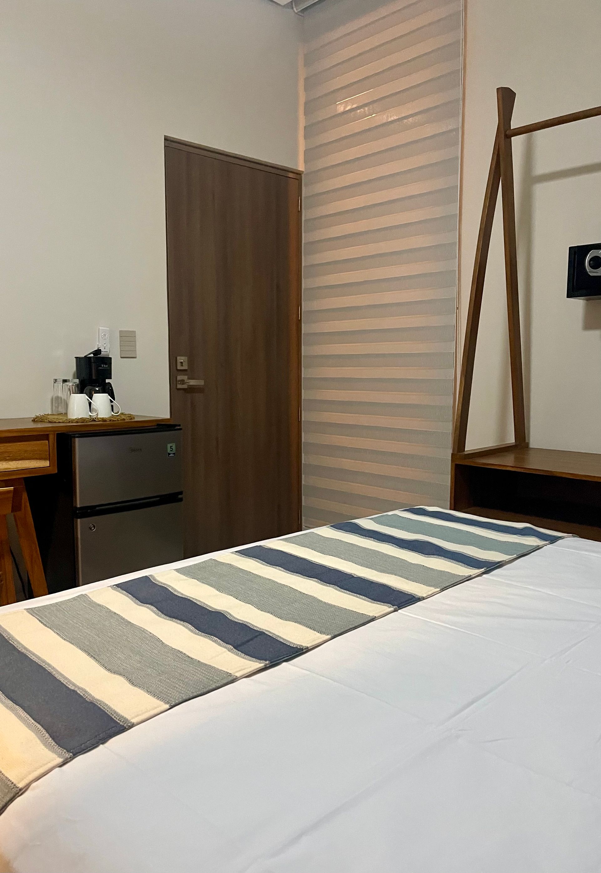 Dormitorio con cama, mininevera, cafetera, puerta y perchero. Detalles en azul claro, blanco y gris.