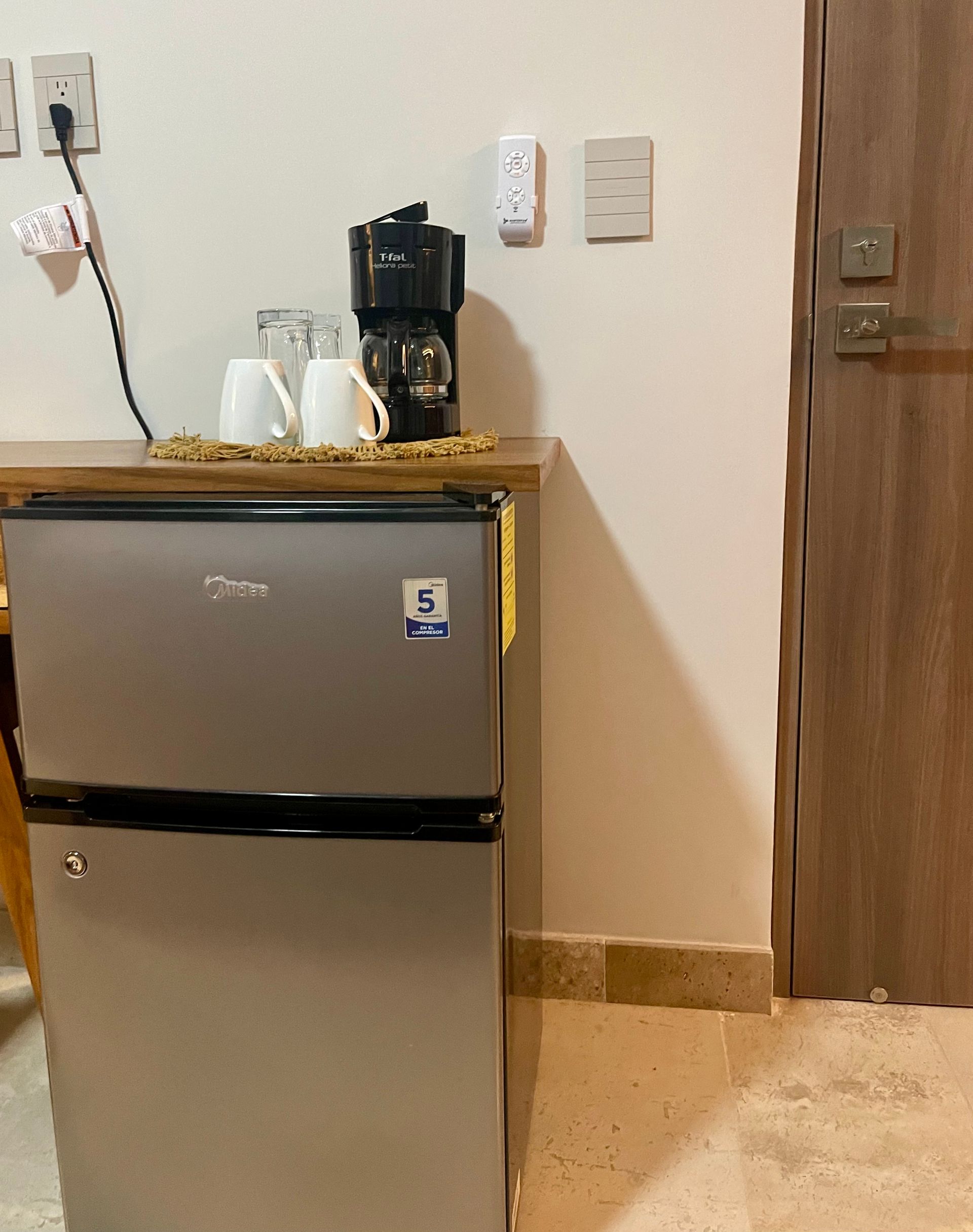 Un pequeño refrigerador con cafetera, tazas y una puerta en una habitación.