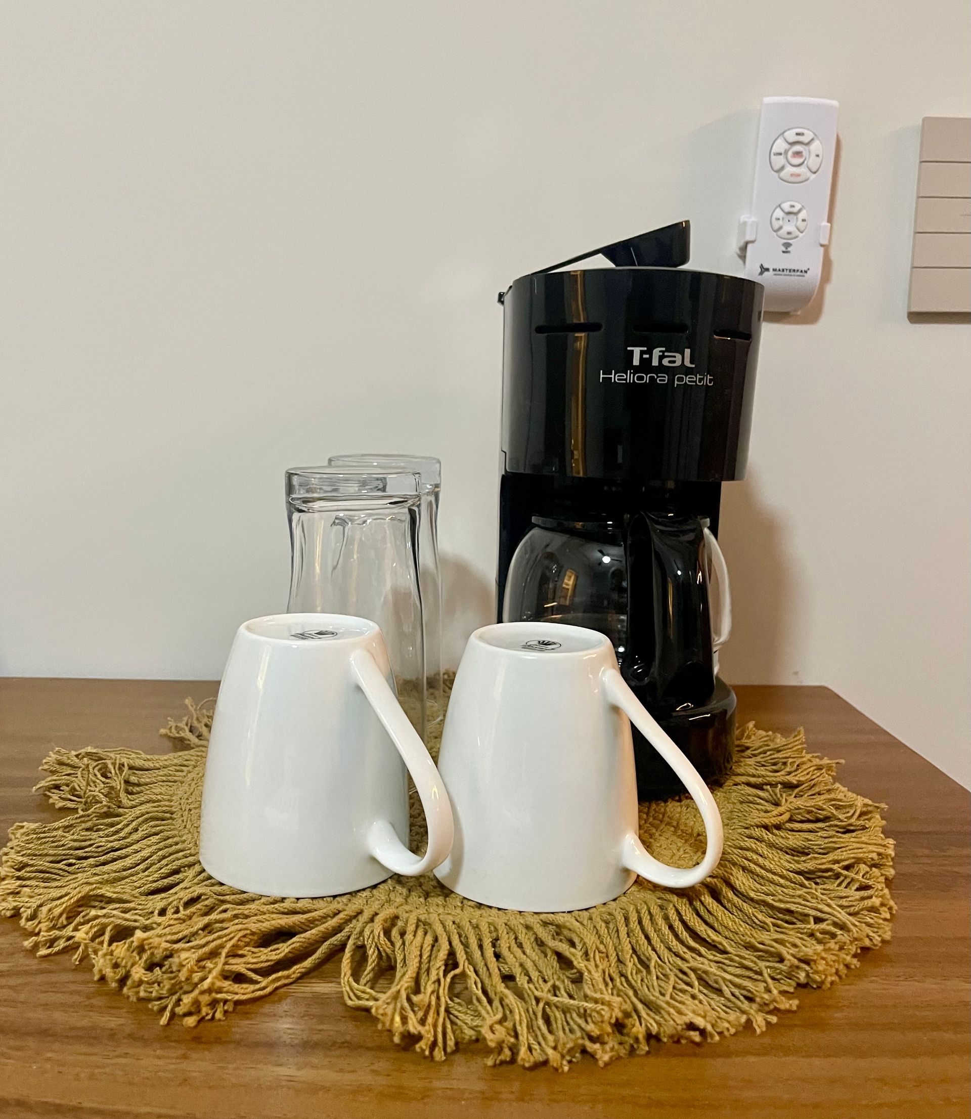 Cafetera con tazas sobre una mesa con tapete con flecos, paredes blancas y una toma de corriente.