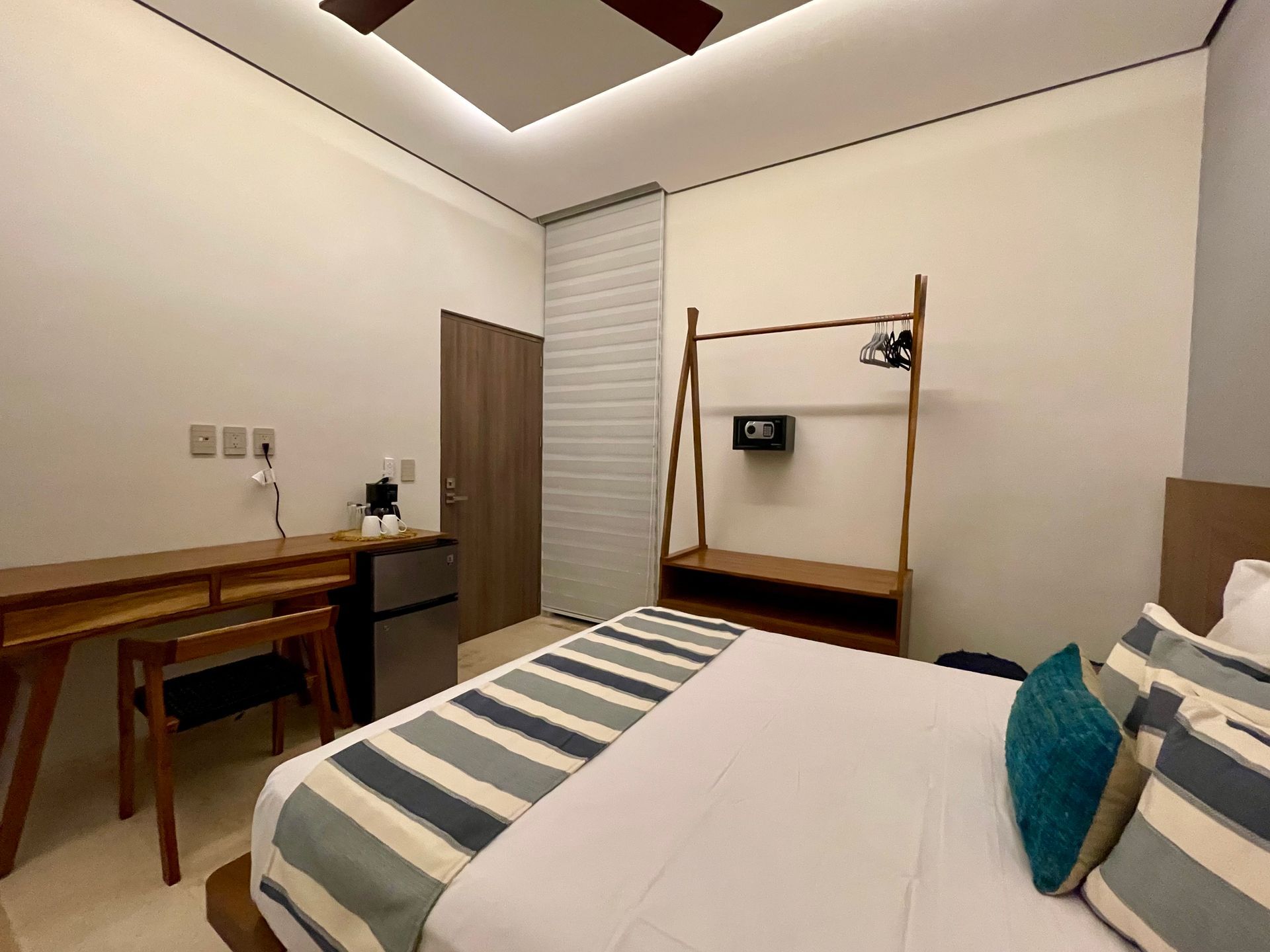 Dormitorio con cama, escritorio, armario, mini-nevera y caja fuerte; paredes color crema y muebles de madera.