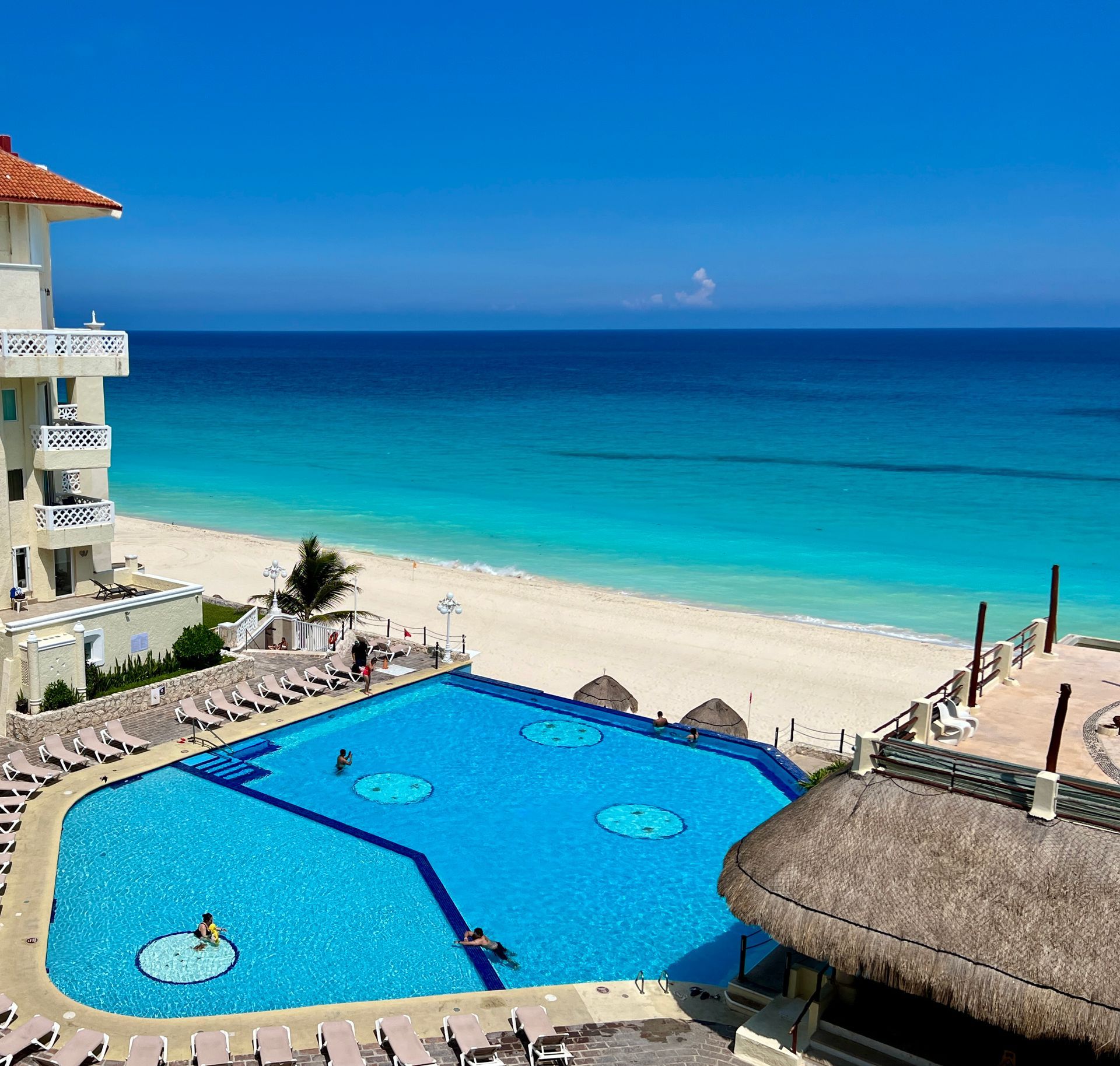 Pool and building overlooking a turquoise ocean and white sand beach on a sunny day.