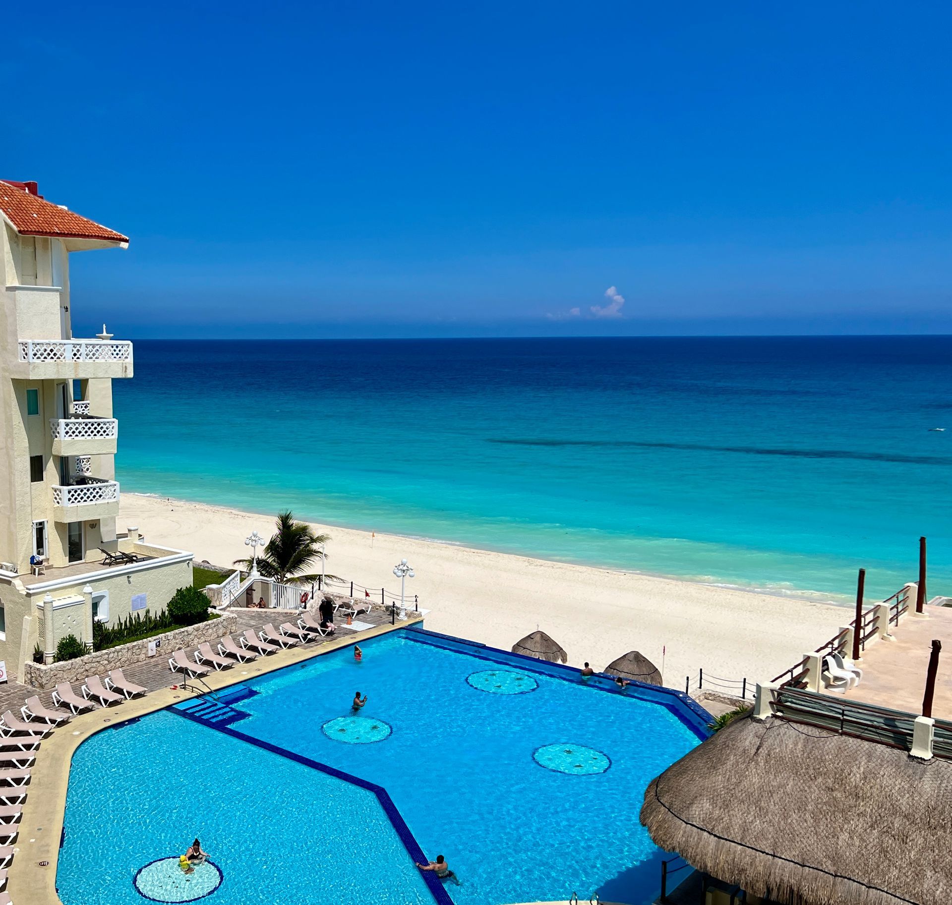 Resort frente al mar con piscina, agua turquesa, arena blanca y cielo azul claro.