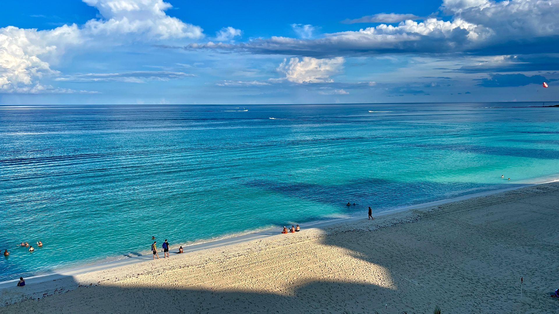 Océano azul con una playa de arena bajo un cielo parcialmente nublado. La gente se relaja en la arena y en el agua.
