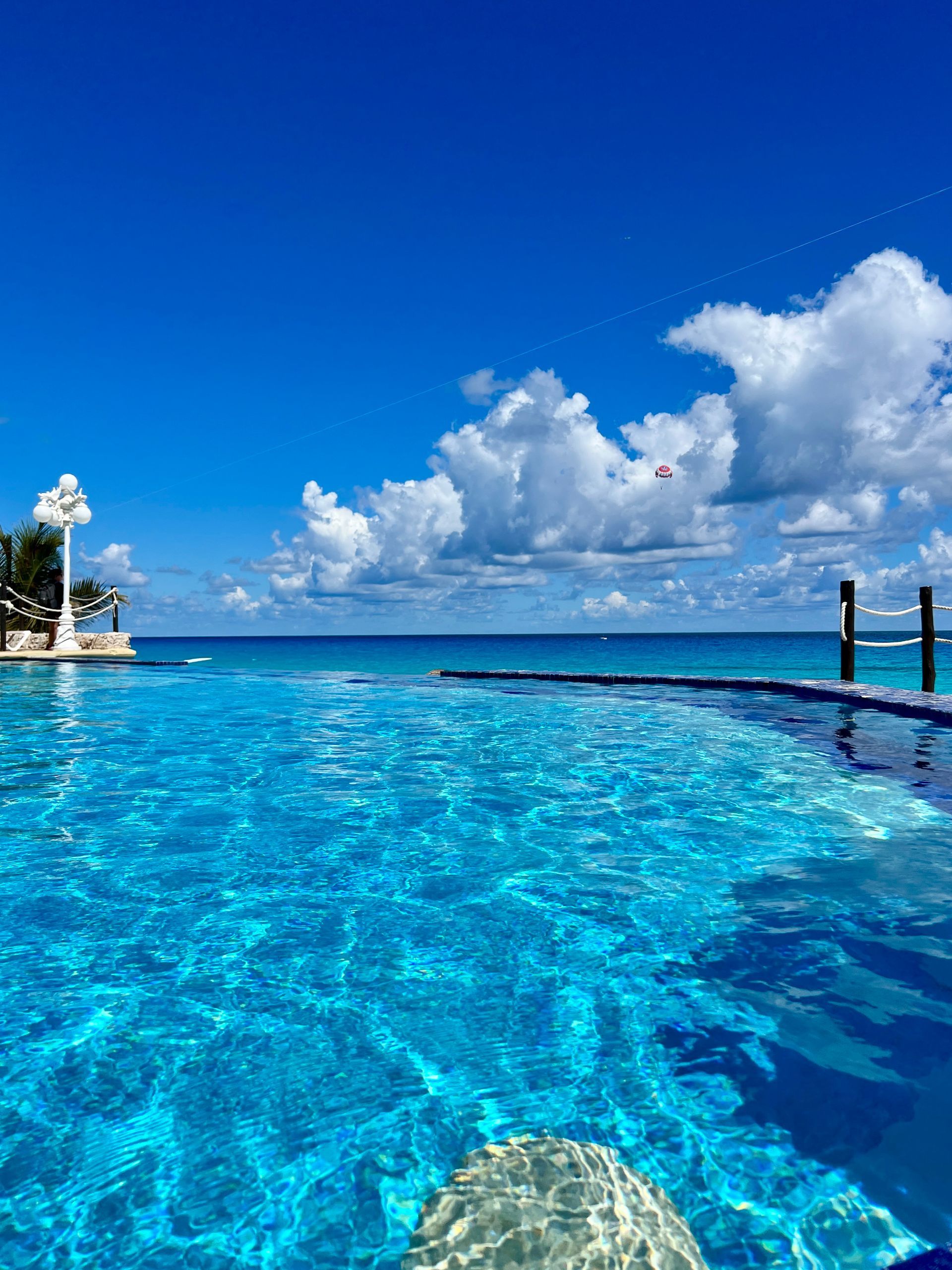 Piscina infinita con vista al océano en un día brillante y soleado; cielo azul claro con nubes blancas esponjosas.