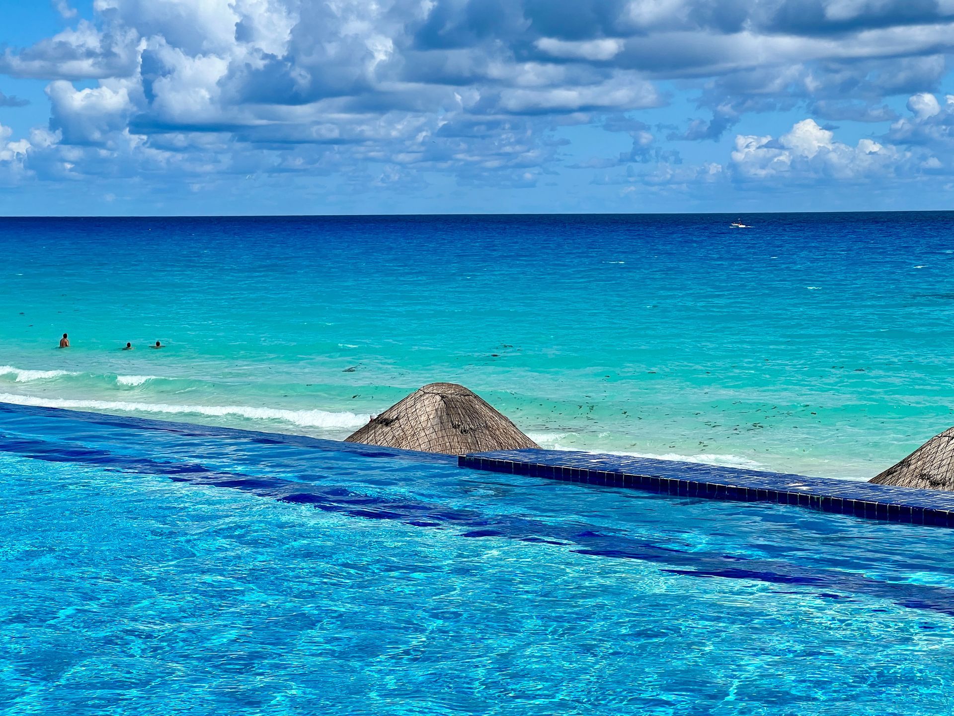 Piscina infinita con vista a un océano turquesa y una playa de arena bajo un cielo parcialmente nublado.