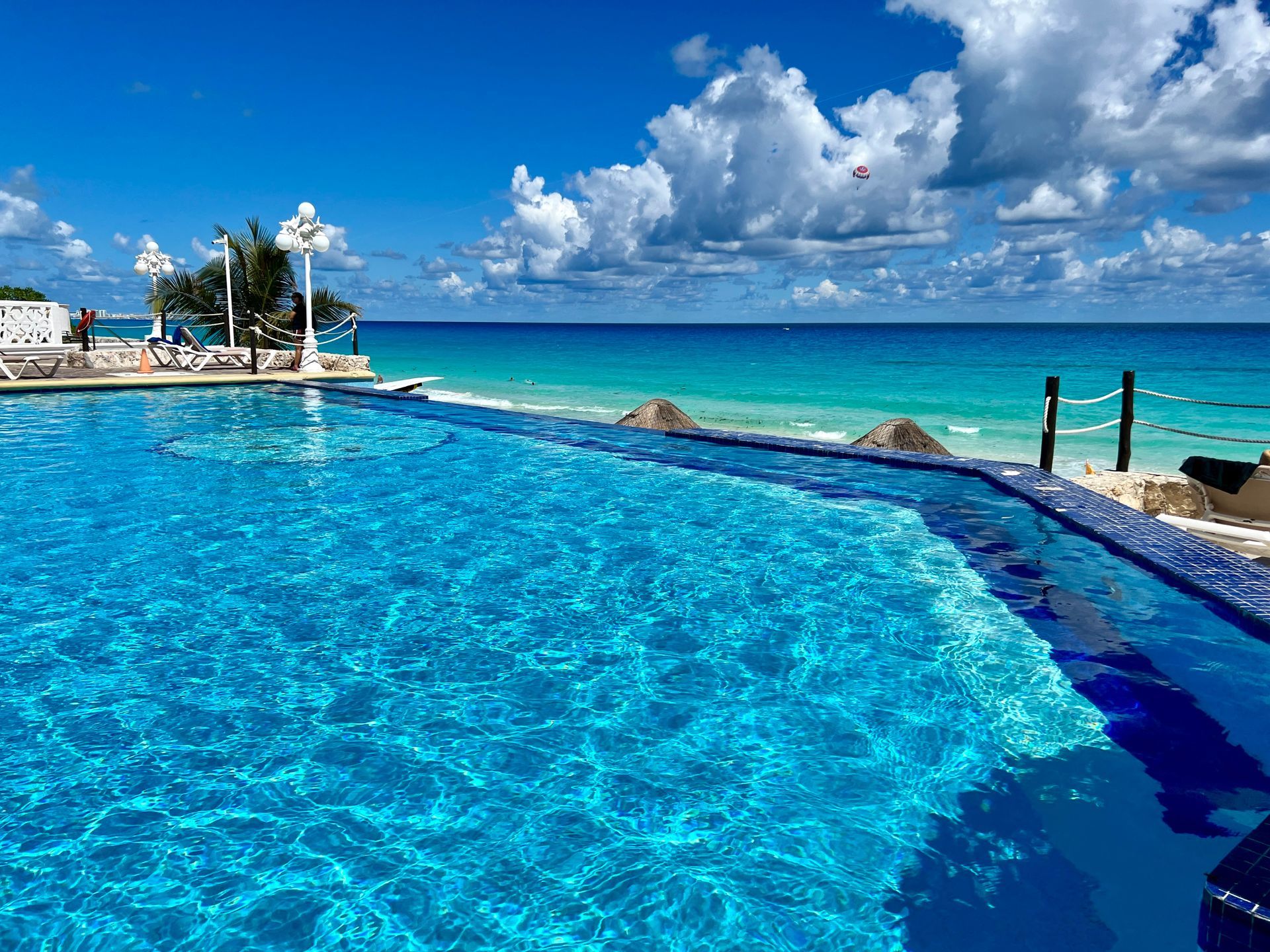 Piscina infinita con vista a un océano turquesa bajo un cielo azul brillante con nubes blancas.