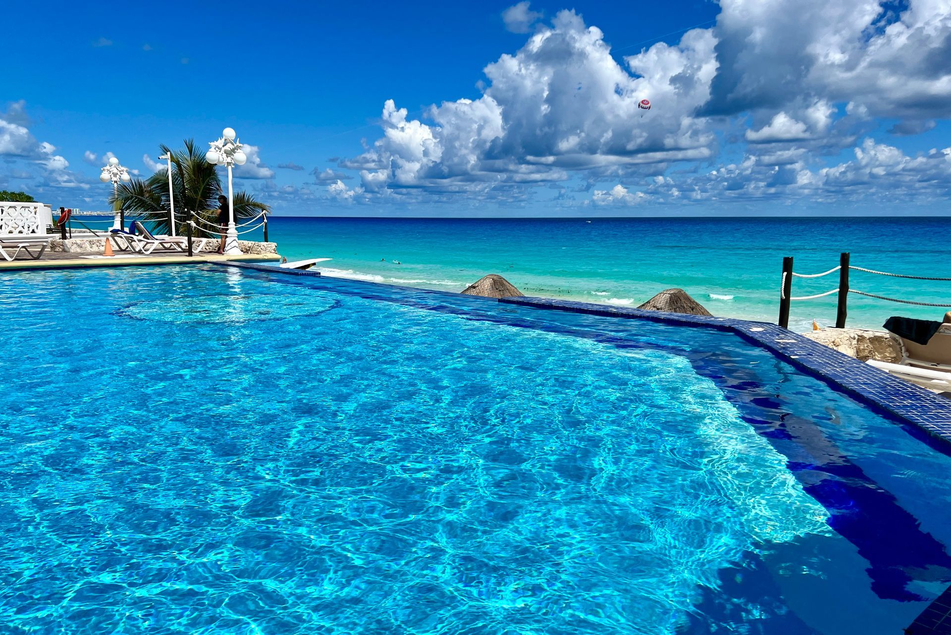 Piscina infinita con vista al océano turquesa bajo un cielo azul con nubes blancas.