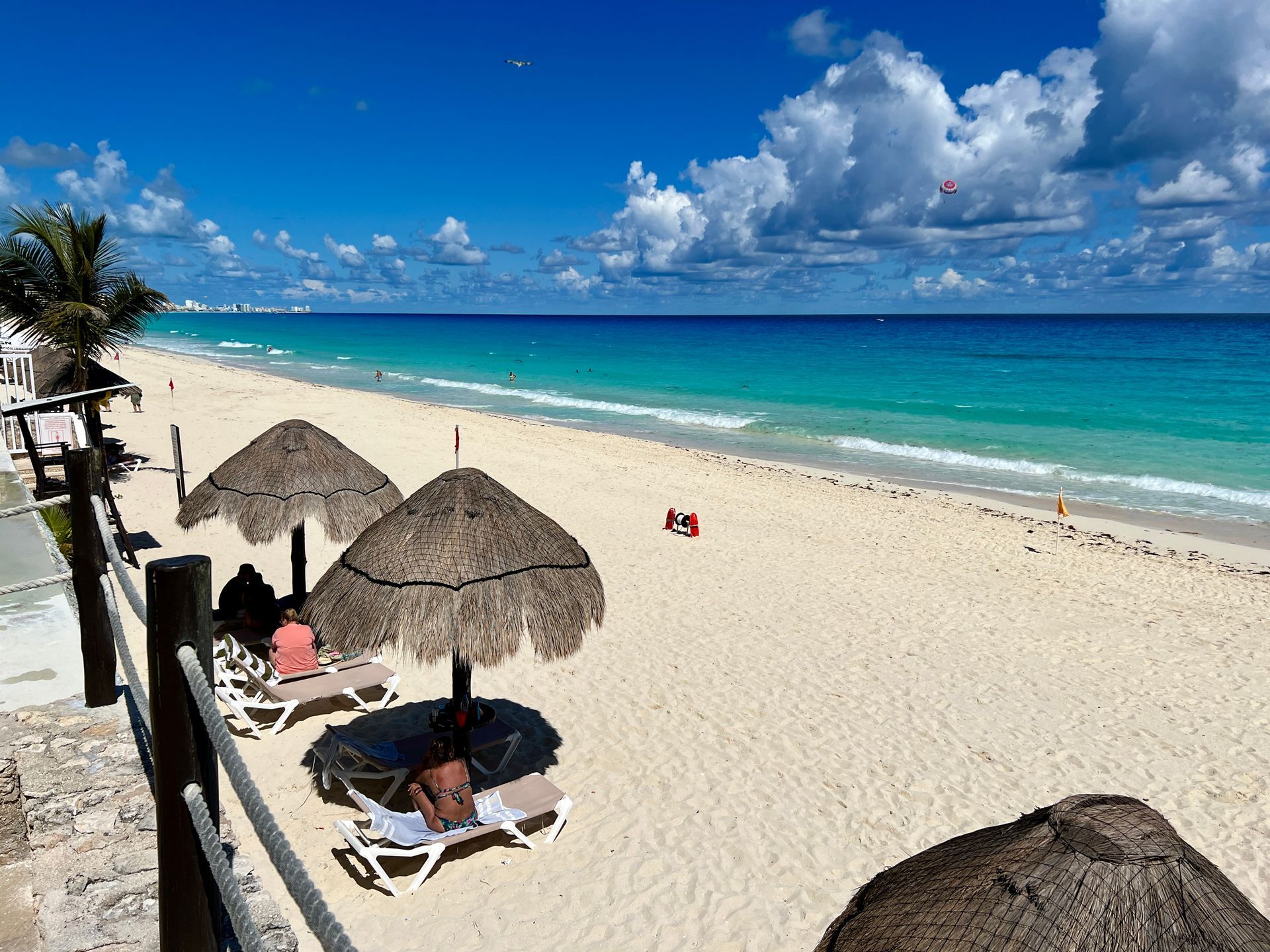 Escena de playa con agua turquesa, arena blanca, sombrillas de paja y un cielo azul brillante.