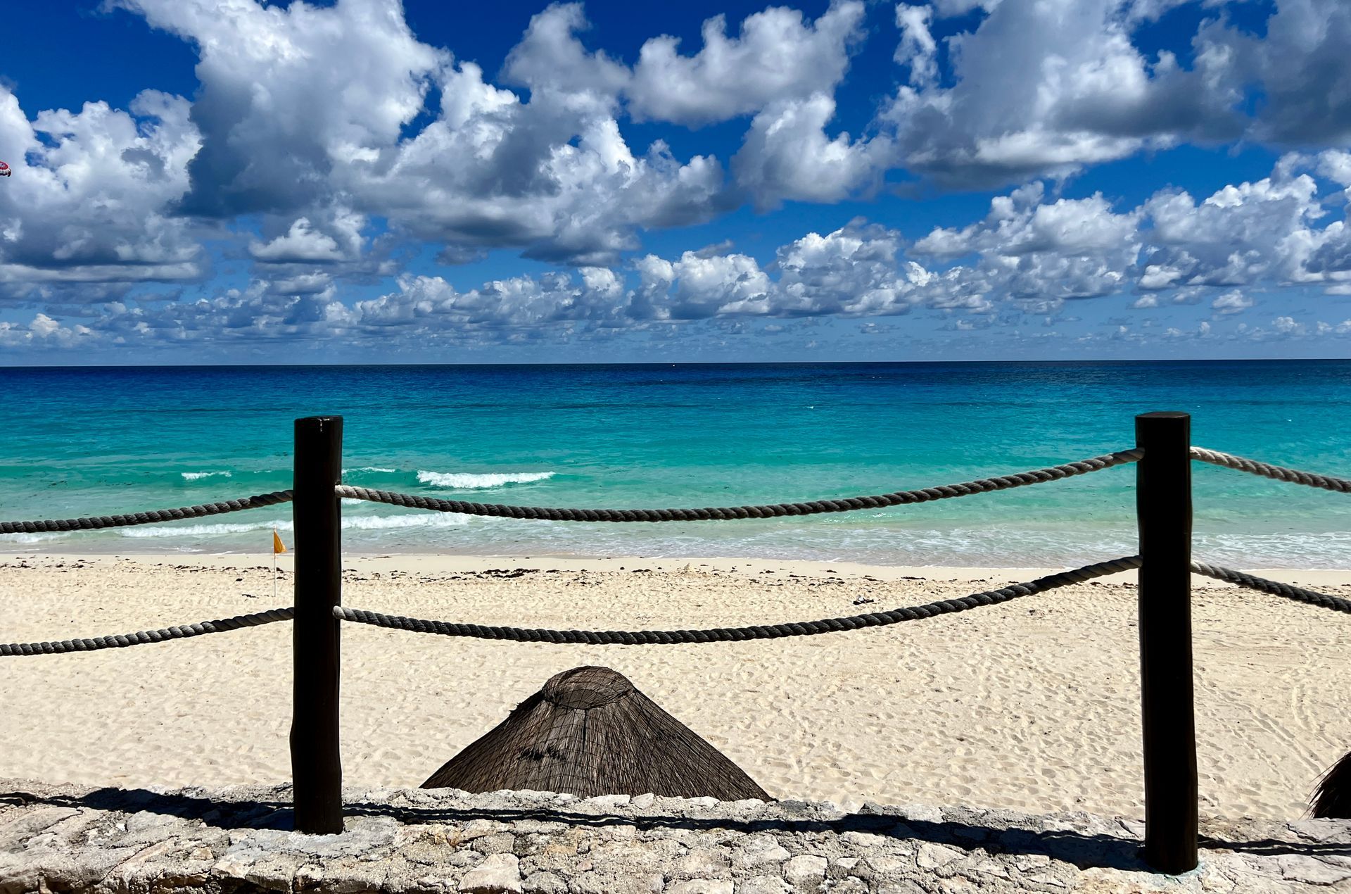 Escena de playa con agua turquesa, arena blanca, cielo azul y nubes blancas. Una barrera de madera en primer plano.