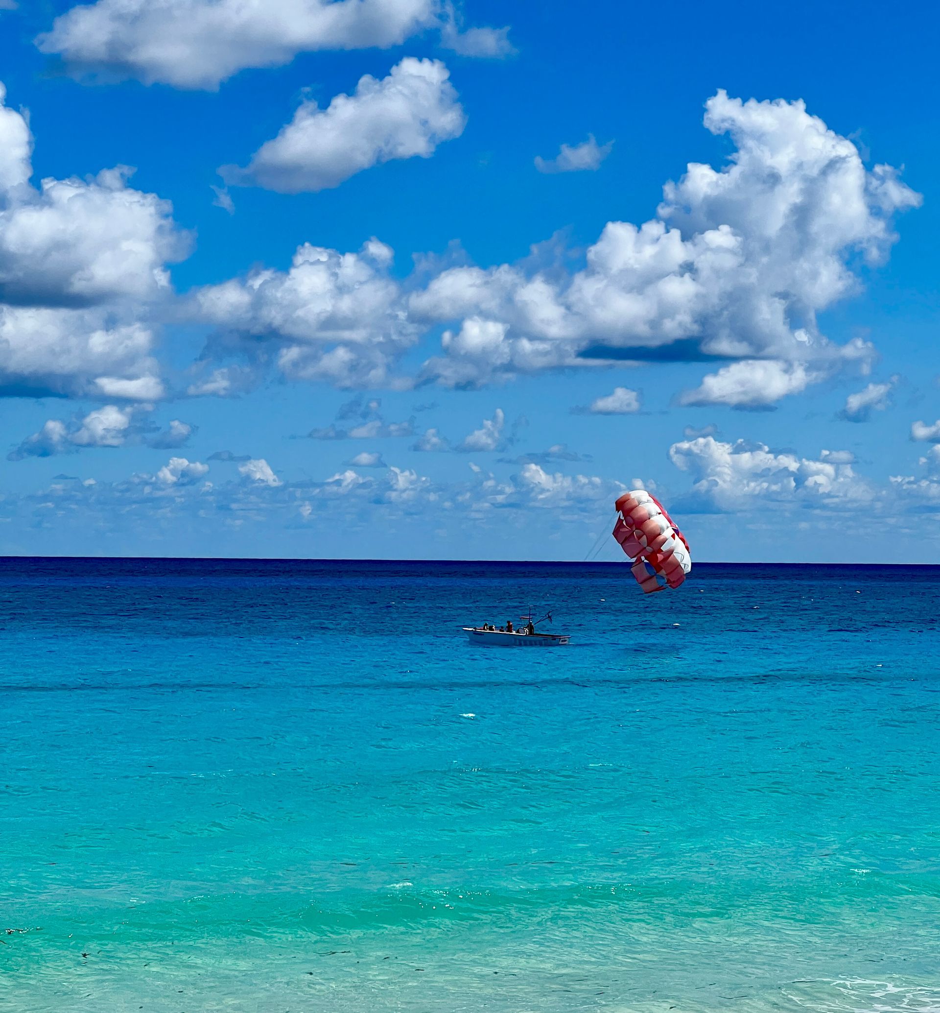Bright blue ocean with white clouds. A parasailer ascends, pulled by a boat in the distance.