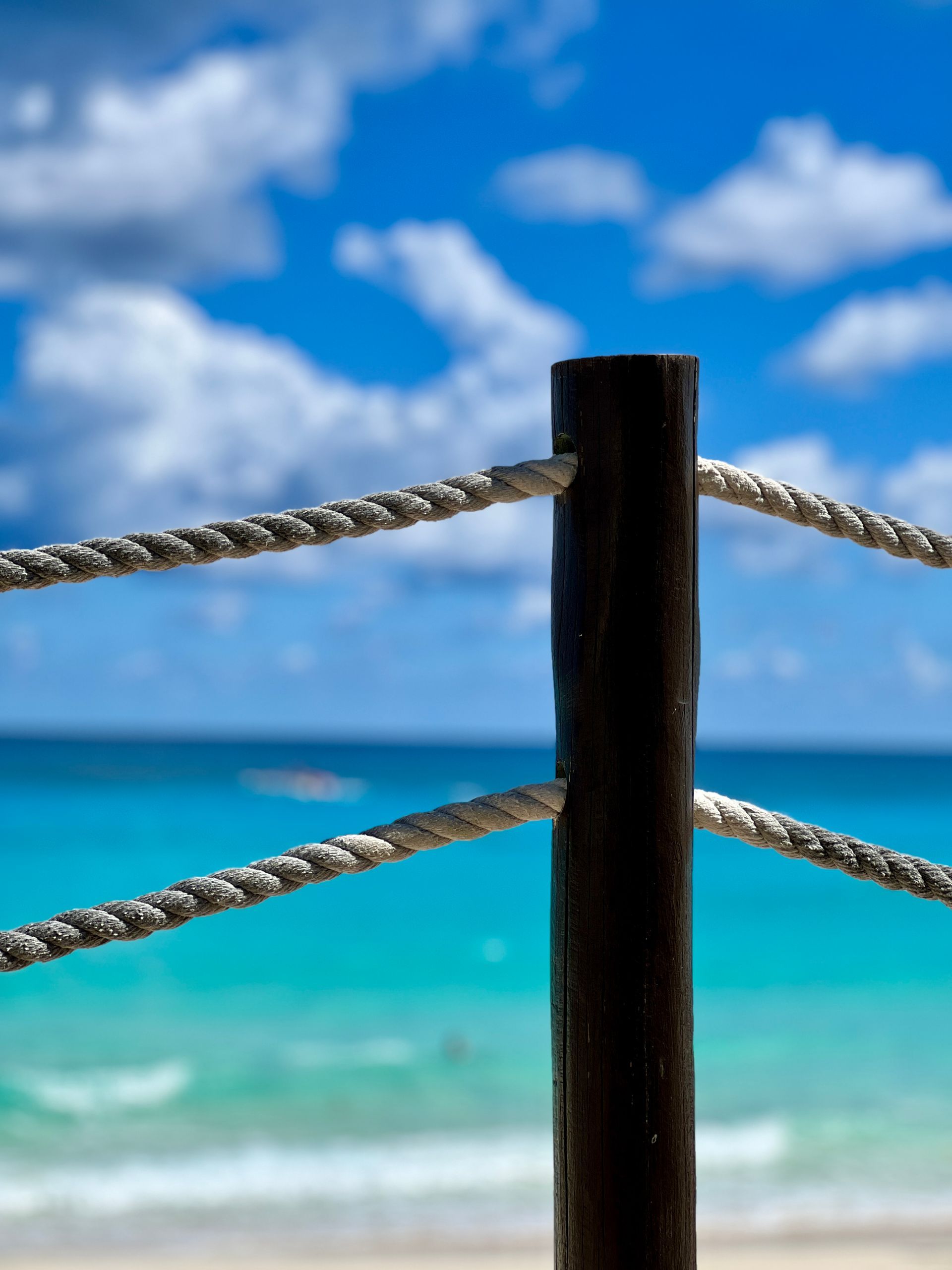 Black post with rope railing overlooking a turquoise ocean and blue sky with clouds.