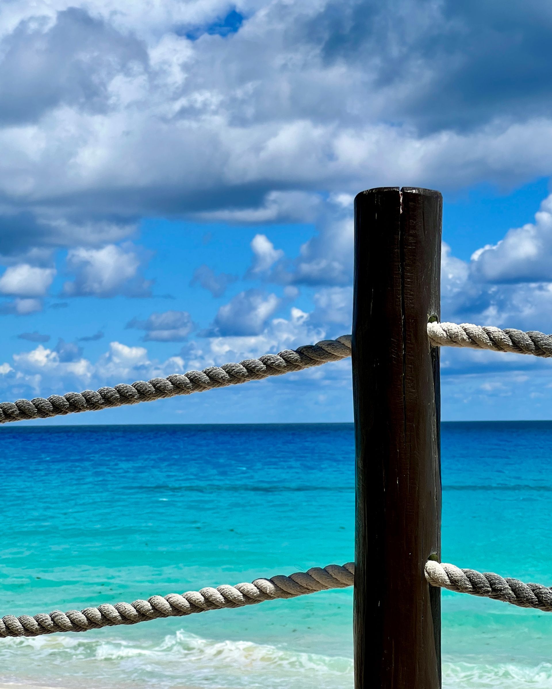Wooden post with rope fence overlooking turquoise ocean and blue sky with clouds.