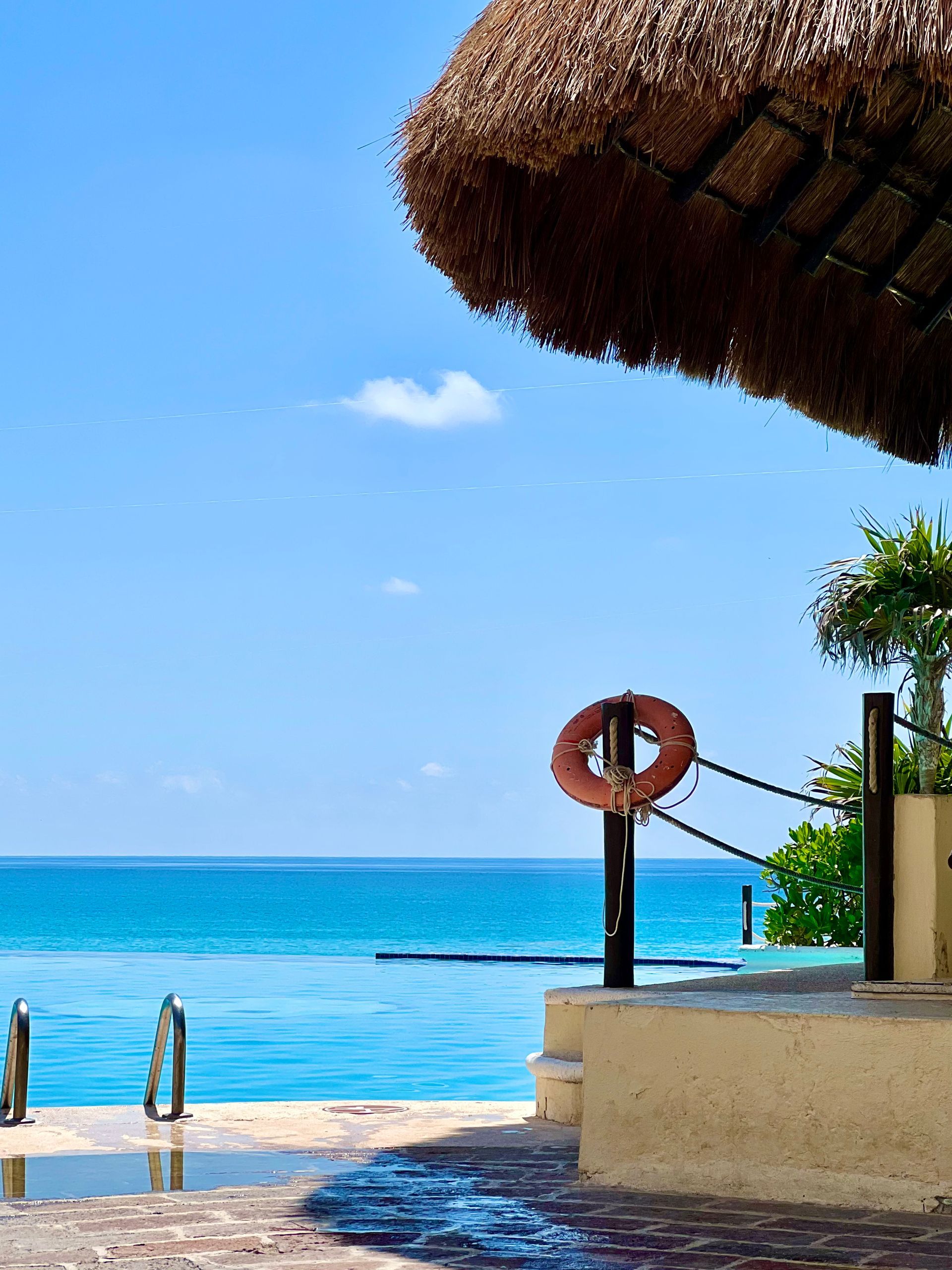 Vista al mar desde una playa con sombrilla de paja, salvavidas y piscina.