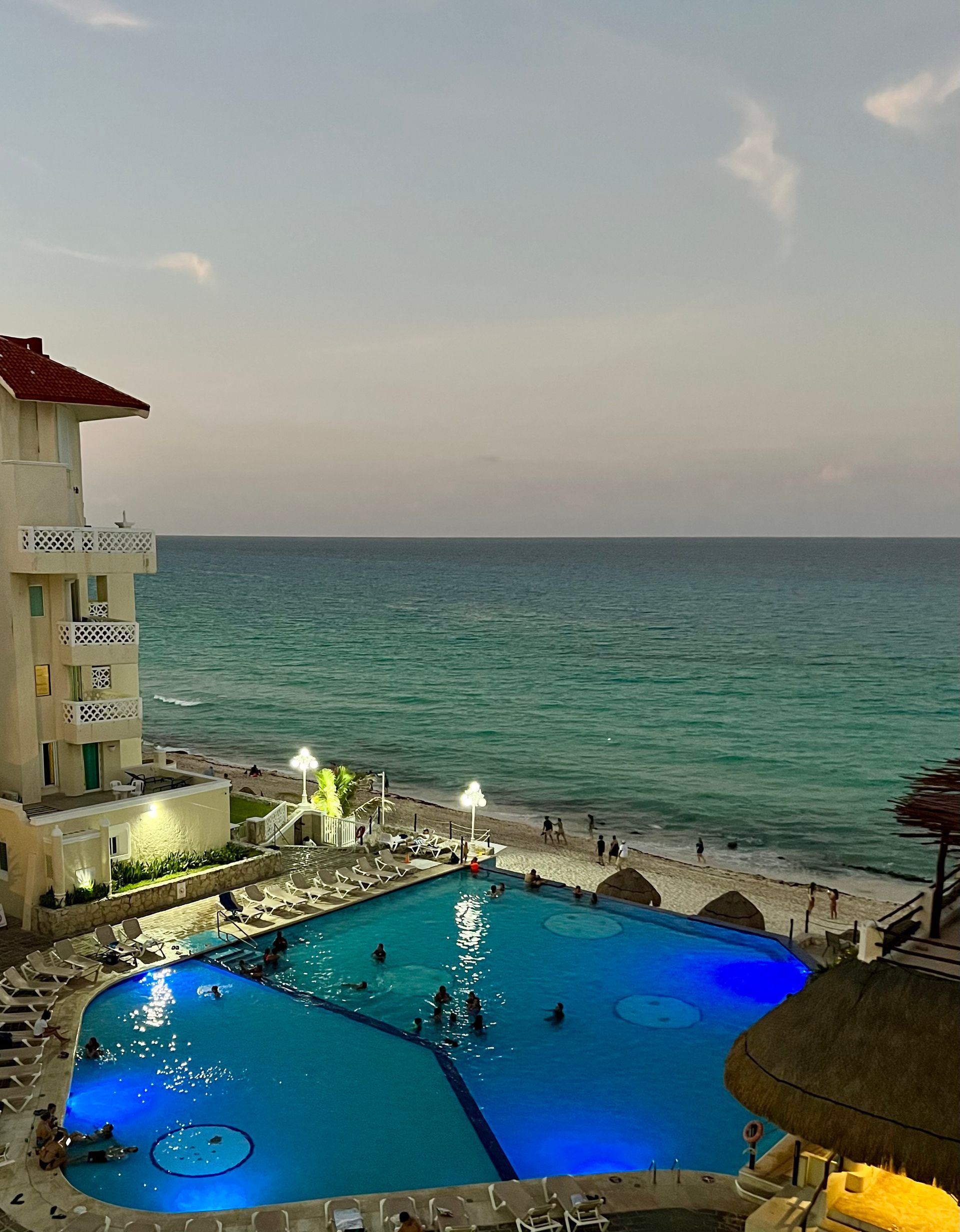 Poolside view of turquoise ocean and a blue lit pool at dusk. A white building is to the left.