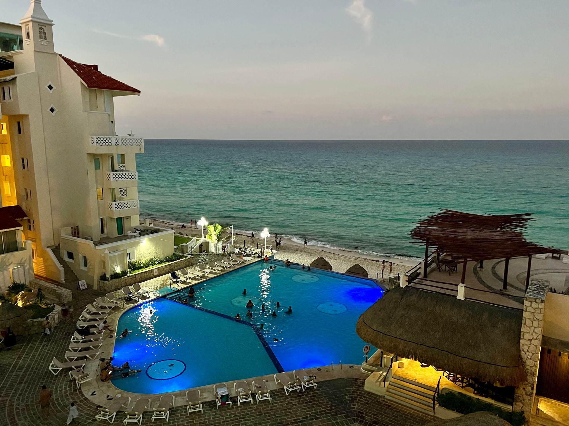 Hotel pool at dusk overlooking the ocean. Lighted pool with people swimming, and a thatch-roofed structure.