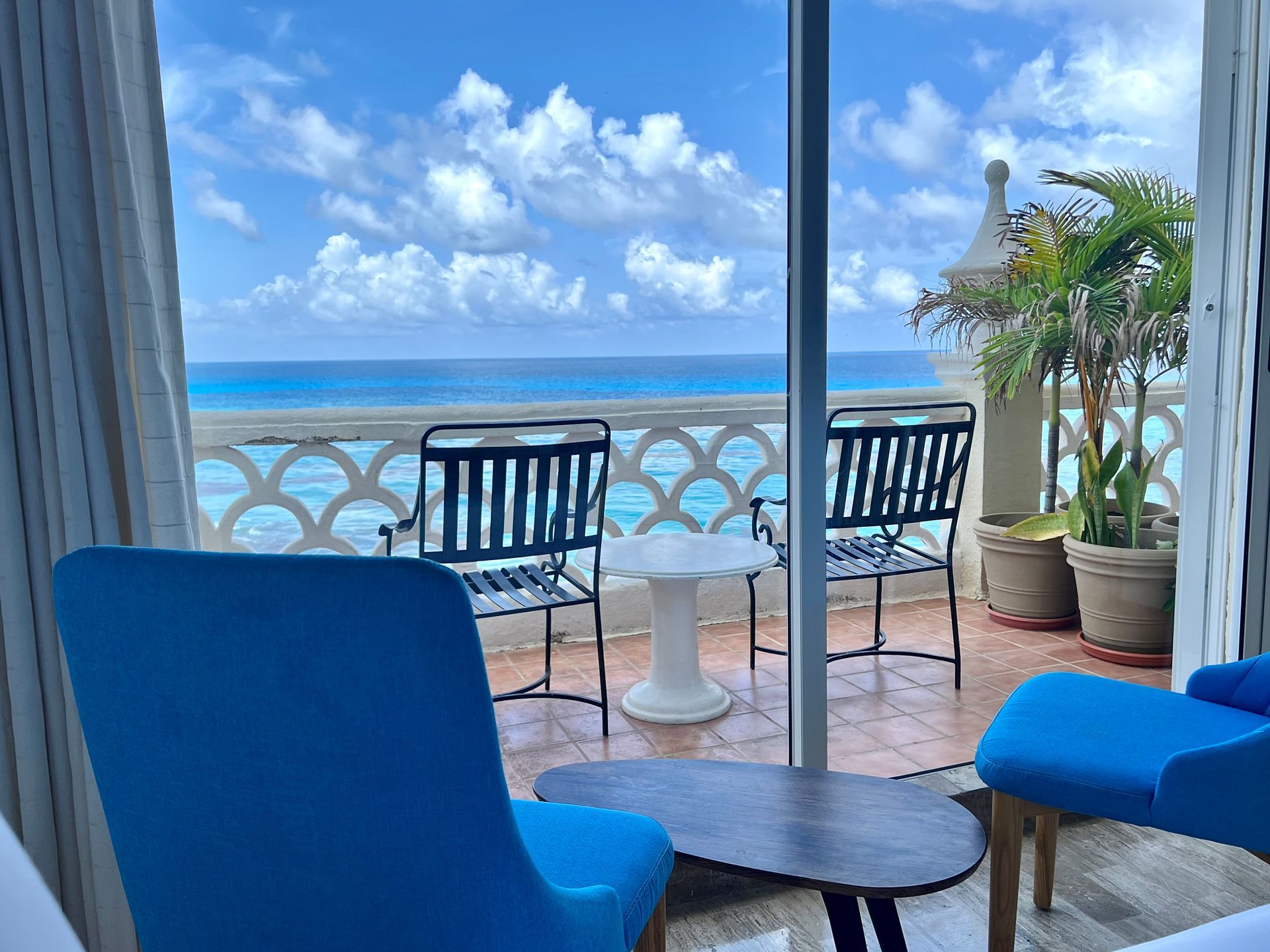 Balcón con sillas azules con vista a un mar turquesa, cielo azul y nubes blancas.