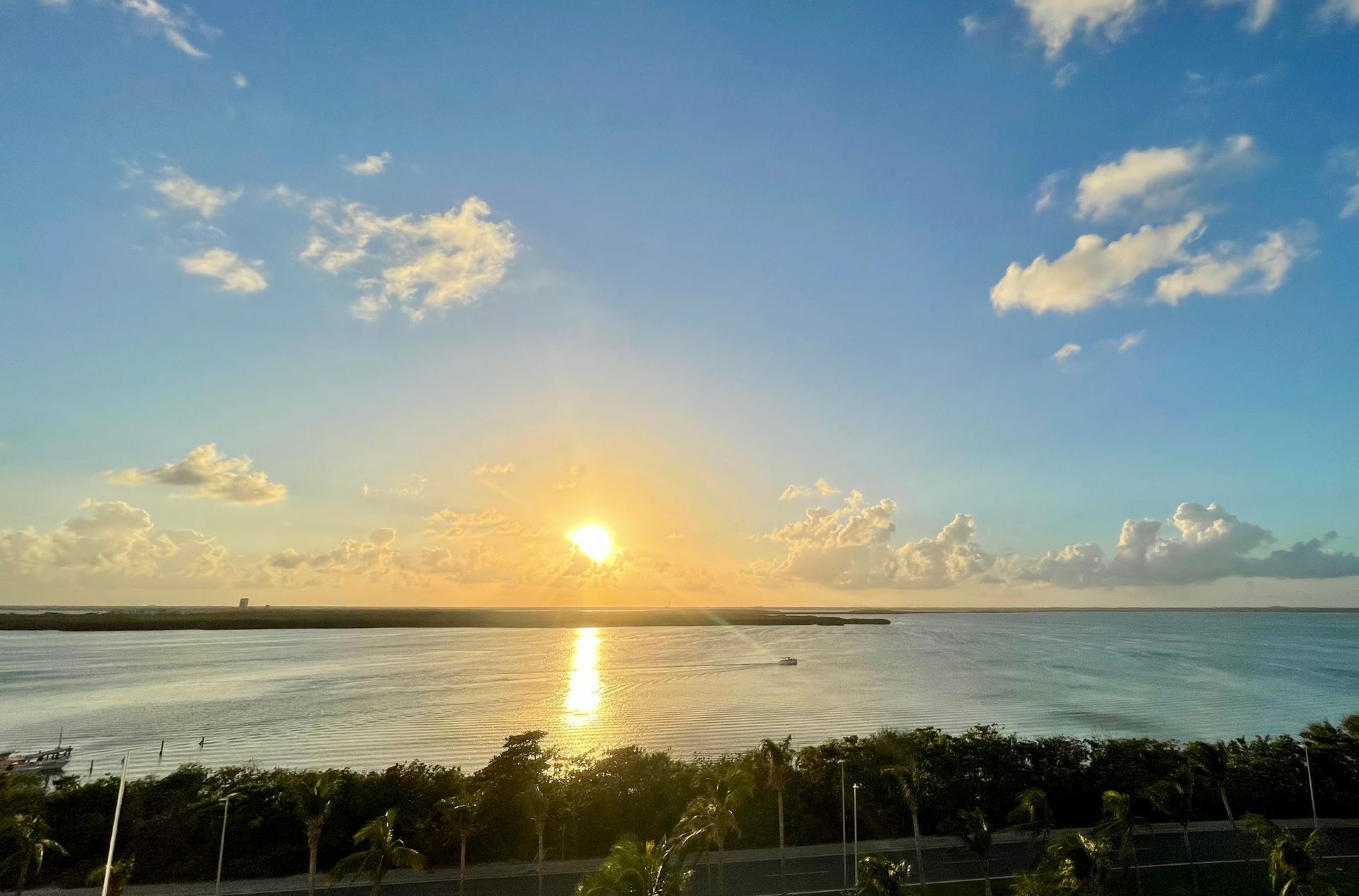 Puesta de sol sobre el agua con sol brillante, cielo azul, nubes y costa verde.