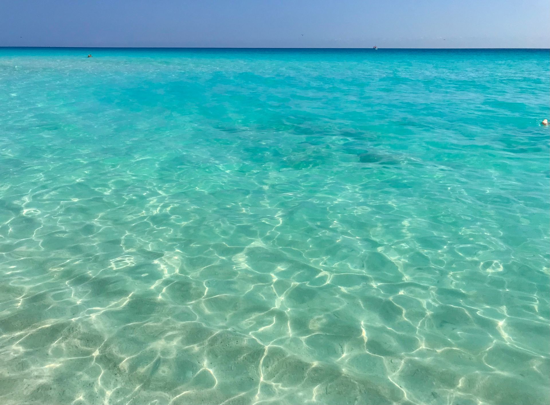 Océano turquesa claro bajo un cielo azul brillante; sol reflejándose en la superficie del agua.
