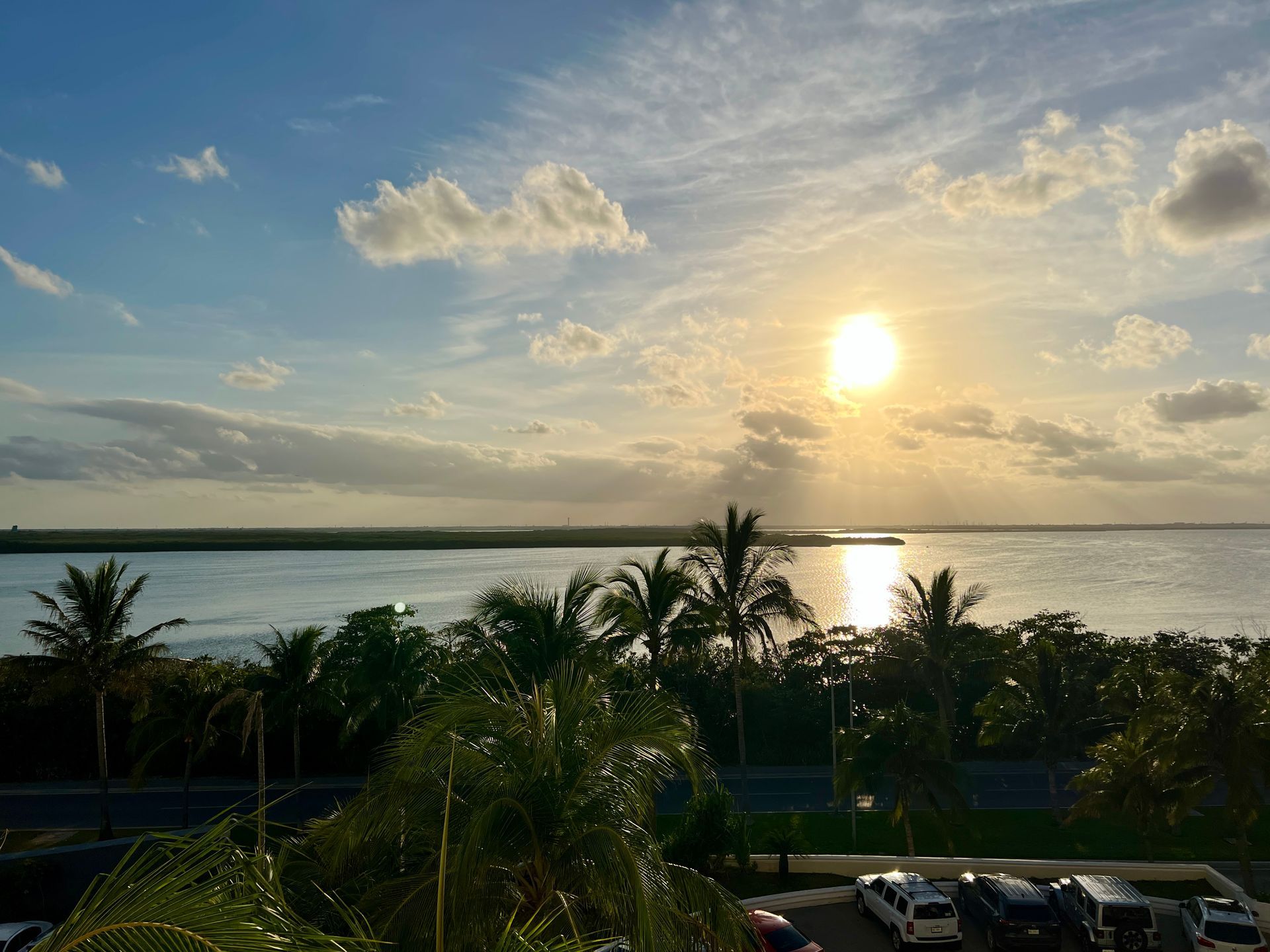 Puesta de sol sobre el agua, palmeras en primer plano, nubes y cielo.