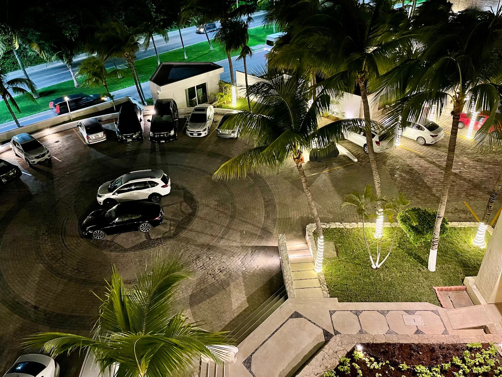 Vista aérea nocturna de un aparcamiento con coches, palmeras y un jardín iluminado.