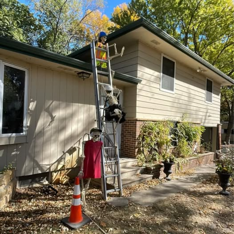 Red Roof Worker on a ladder cleaning gutters beside a beige house, with an orange cone and red safety vest below.
