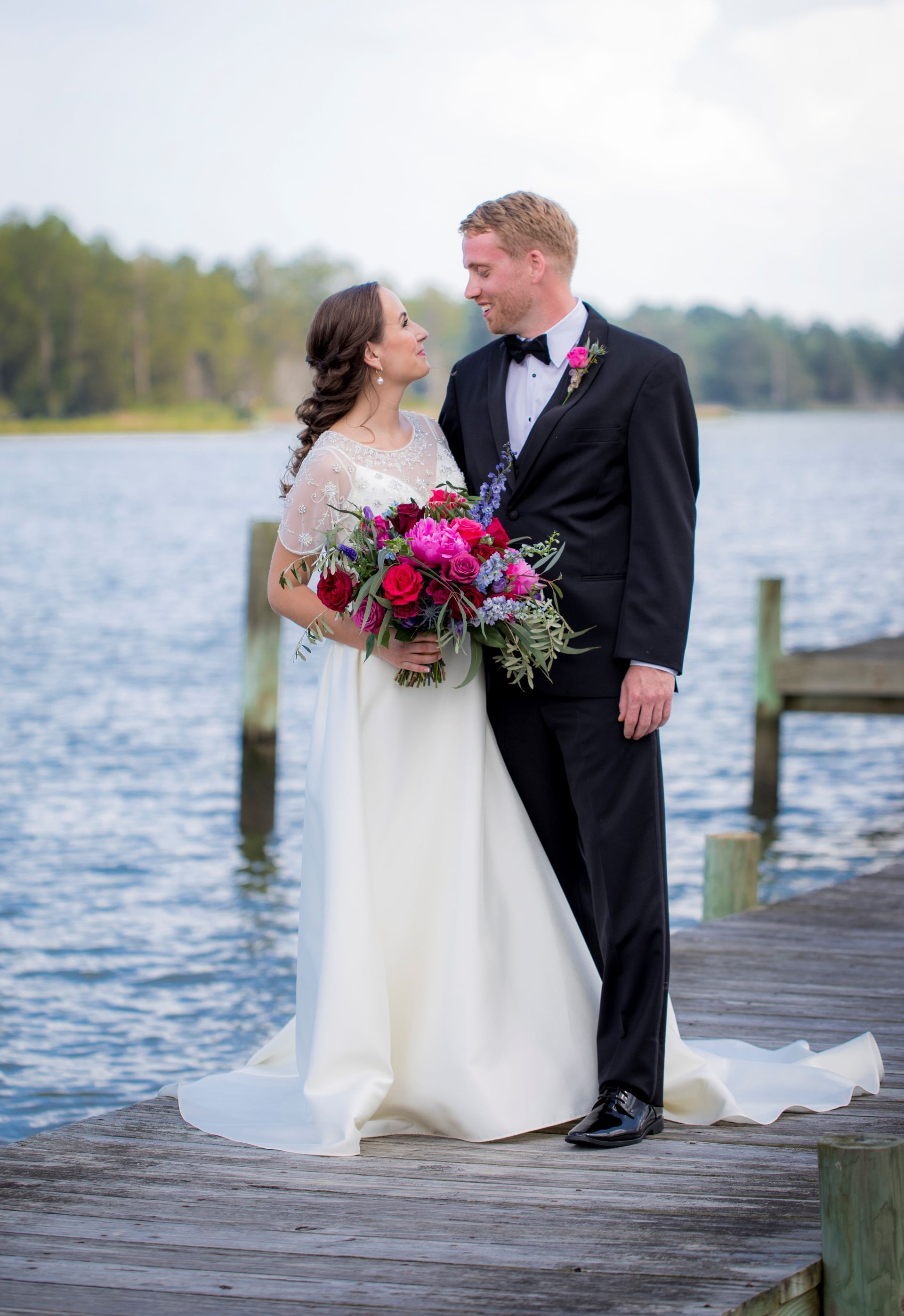 A bride and groom are posing for a picture on a dock next to a lake.