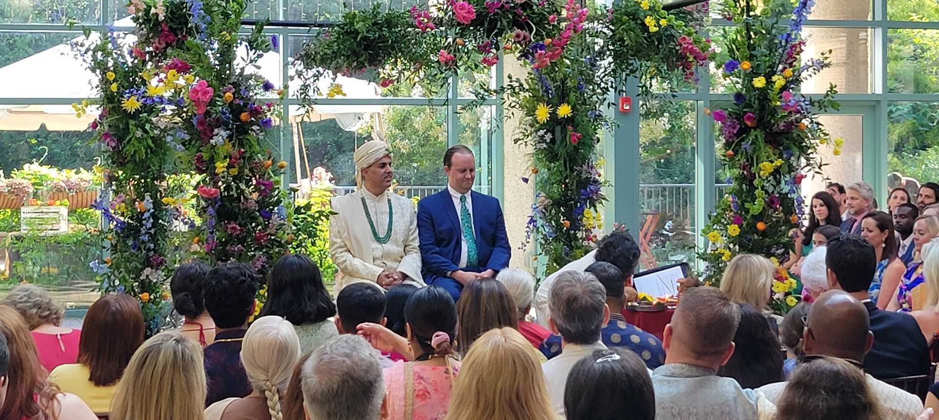 A group of people are sitting in front of a floral arch at a wedding ceremony.