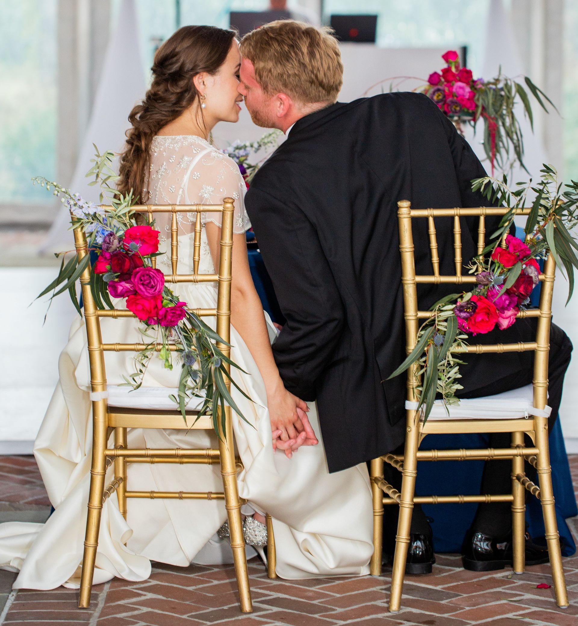 A bride and groom kissing while sitting in chairs decorated with flowers