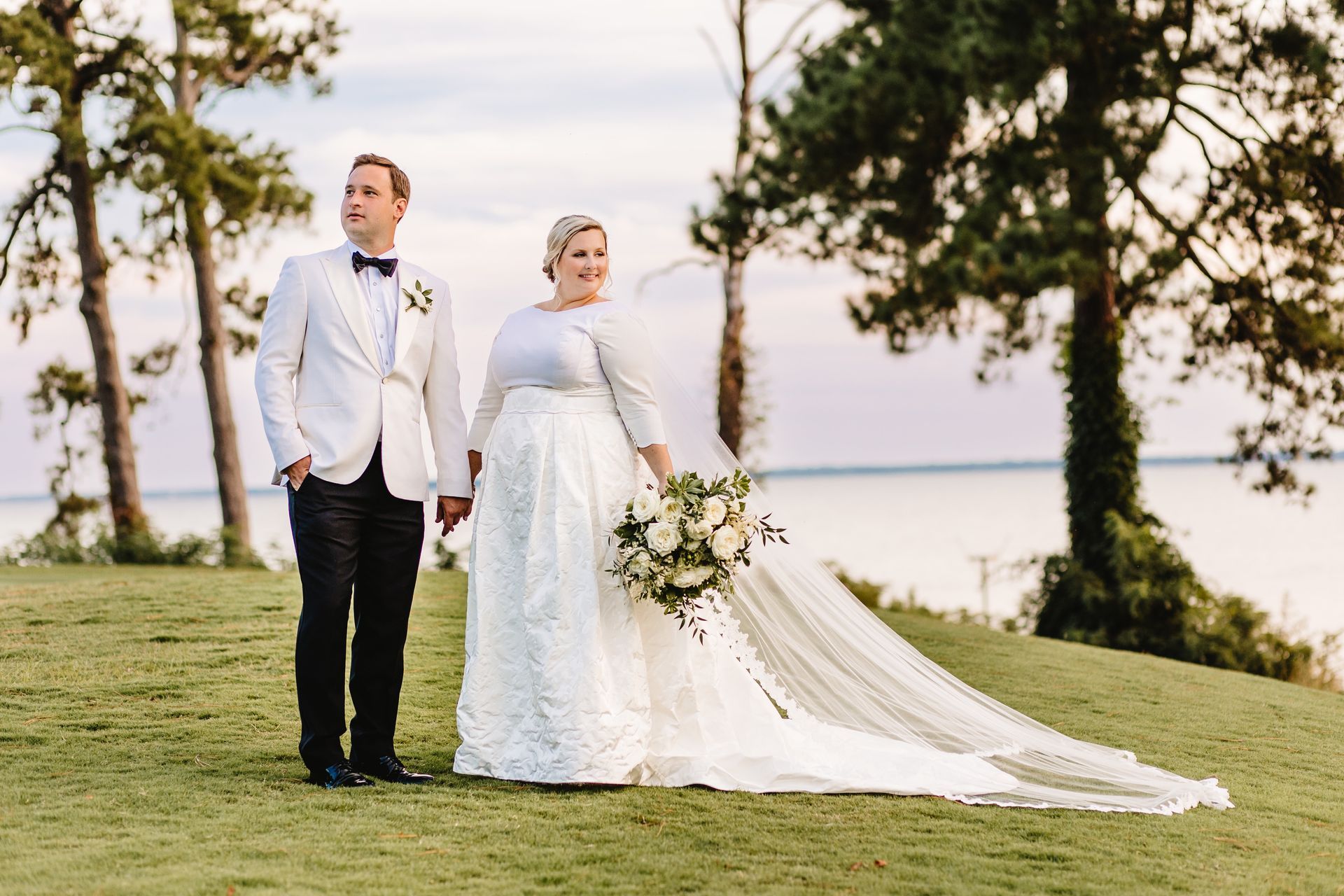 A bride and groom are standing on top of a grassy hill holding hands.