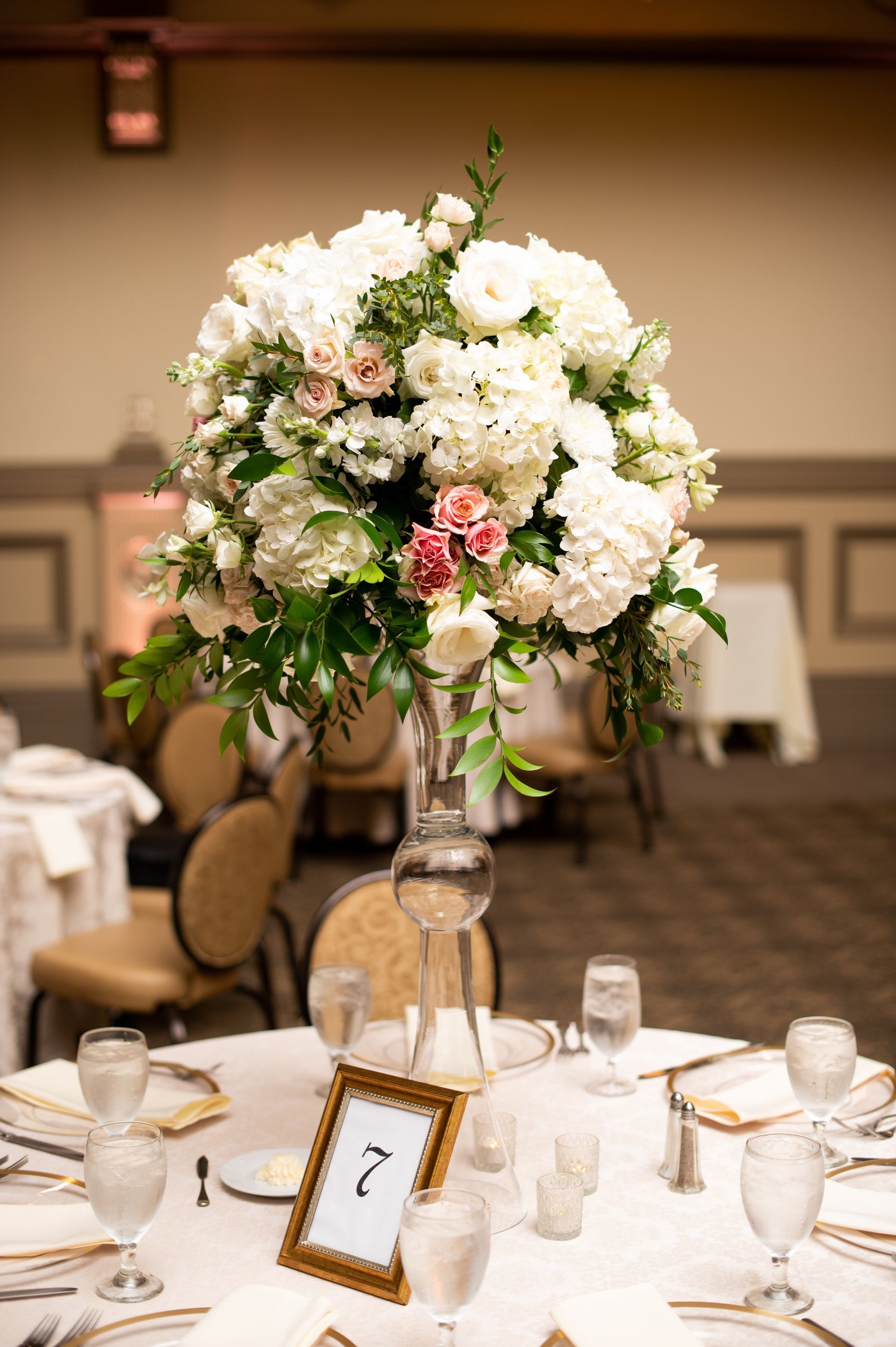 A tall vase filled with white flowers is on a table at a wedding reception.