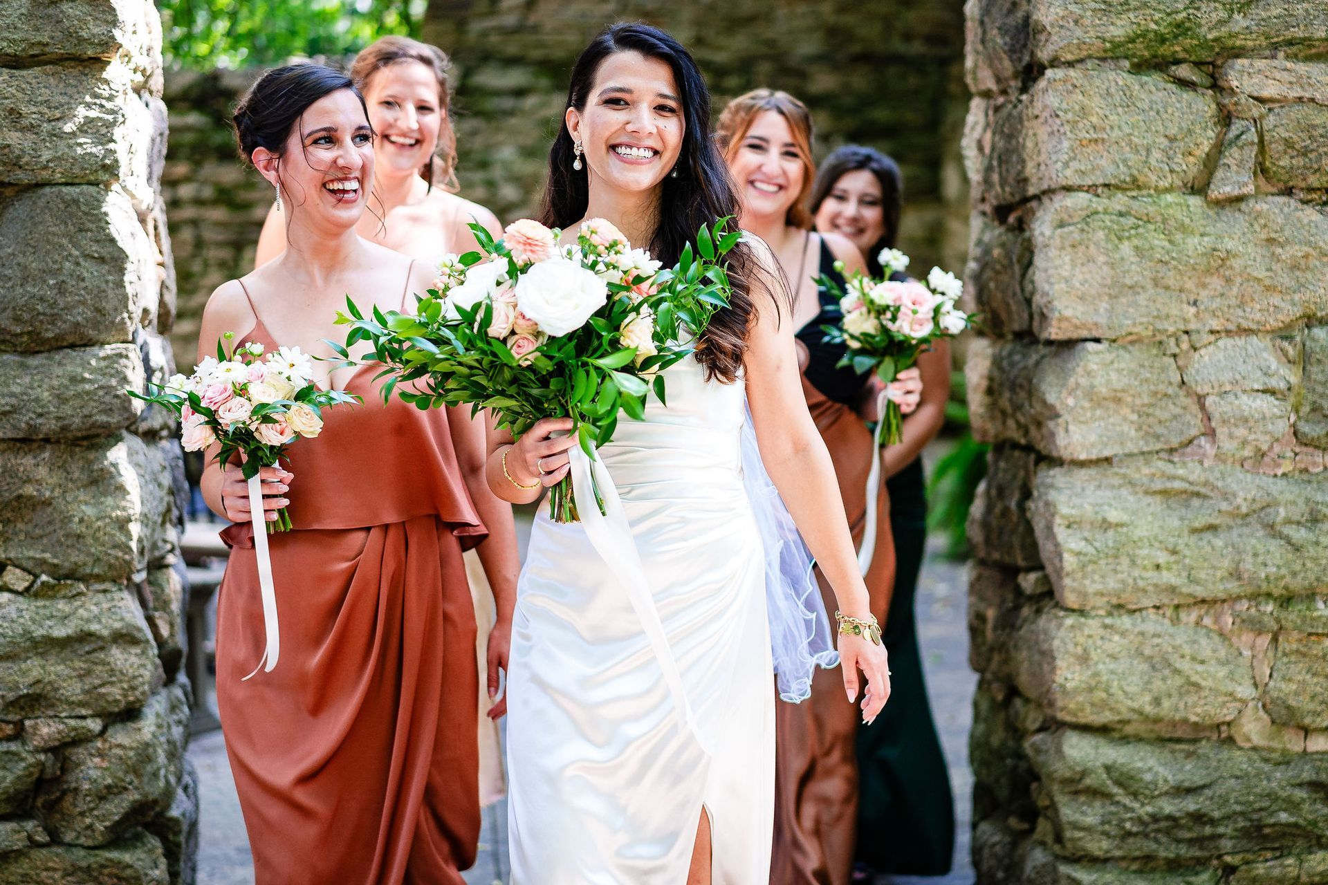A bride and her bridesmaids are walking through a stone archway.