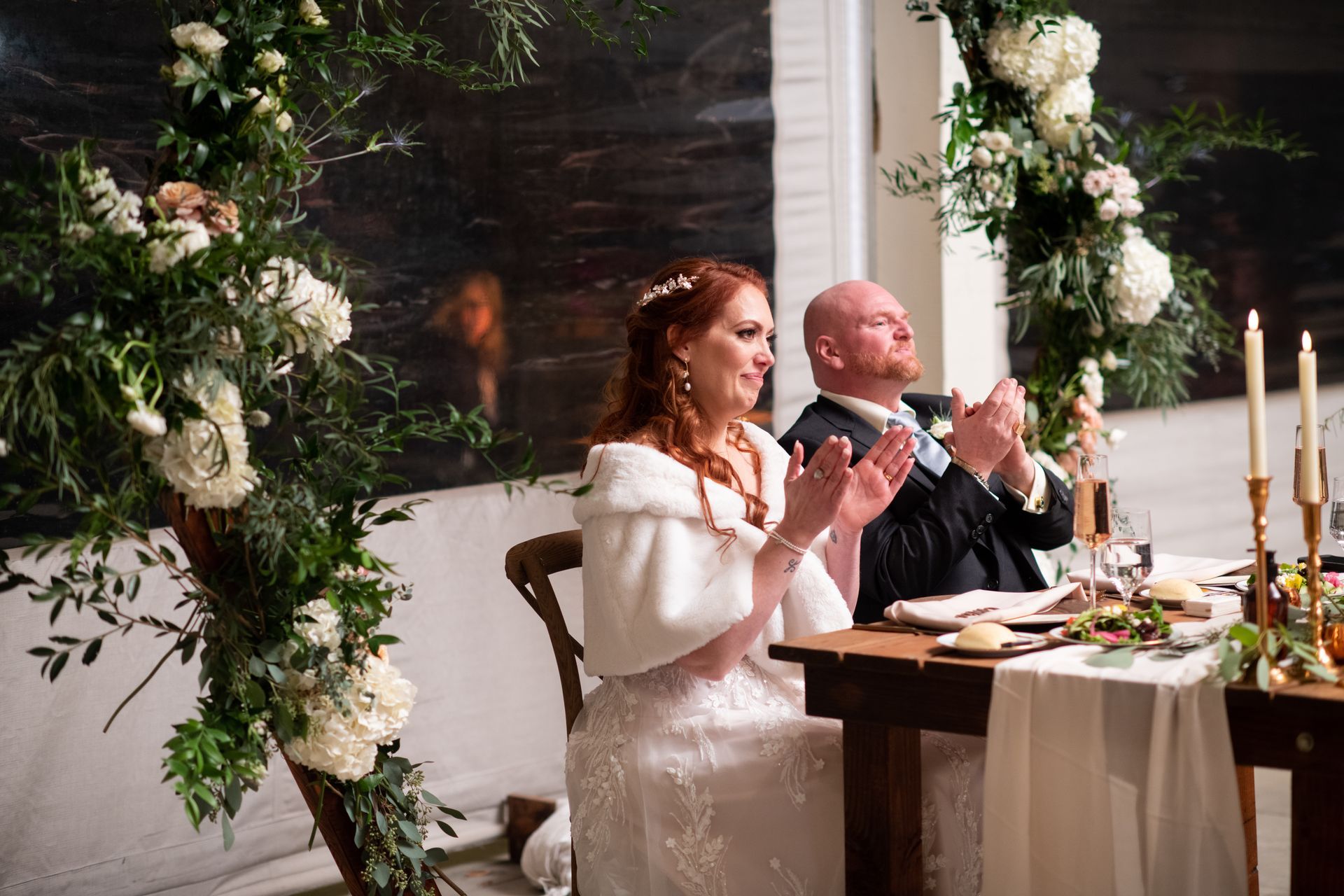 A bride and groom are sitting at a table applauding at their wedding reception.