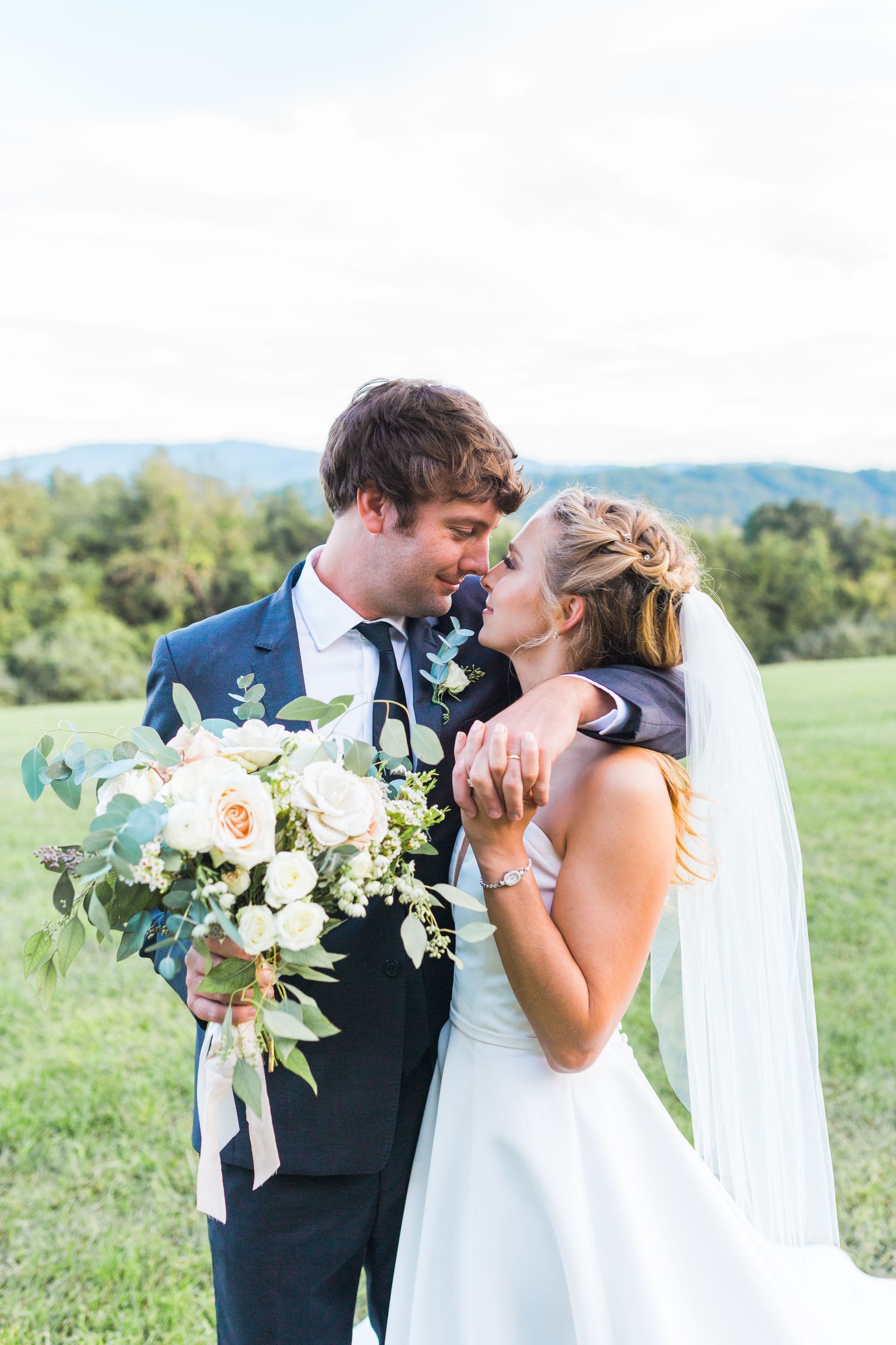 A bride and groom are kissing in a field while the bride is holding a bouquet of flowers.