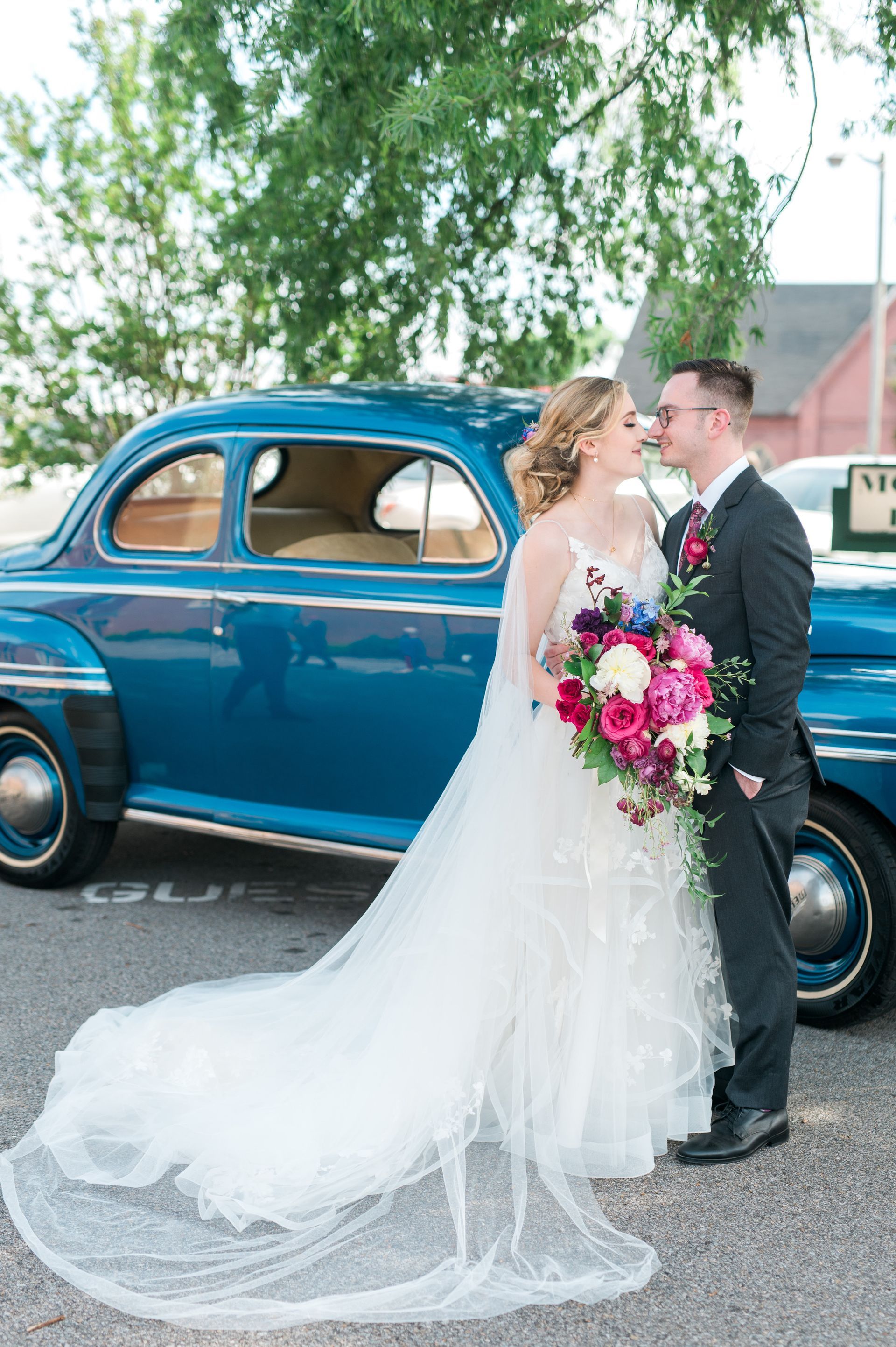 A bride and groom are posing for a picture in front of a blue car.